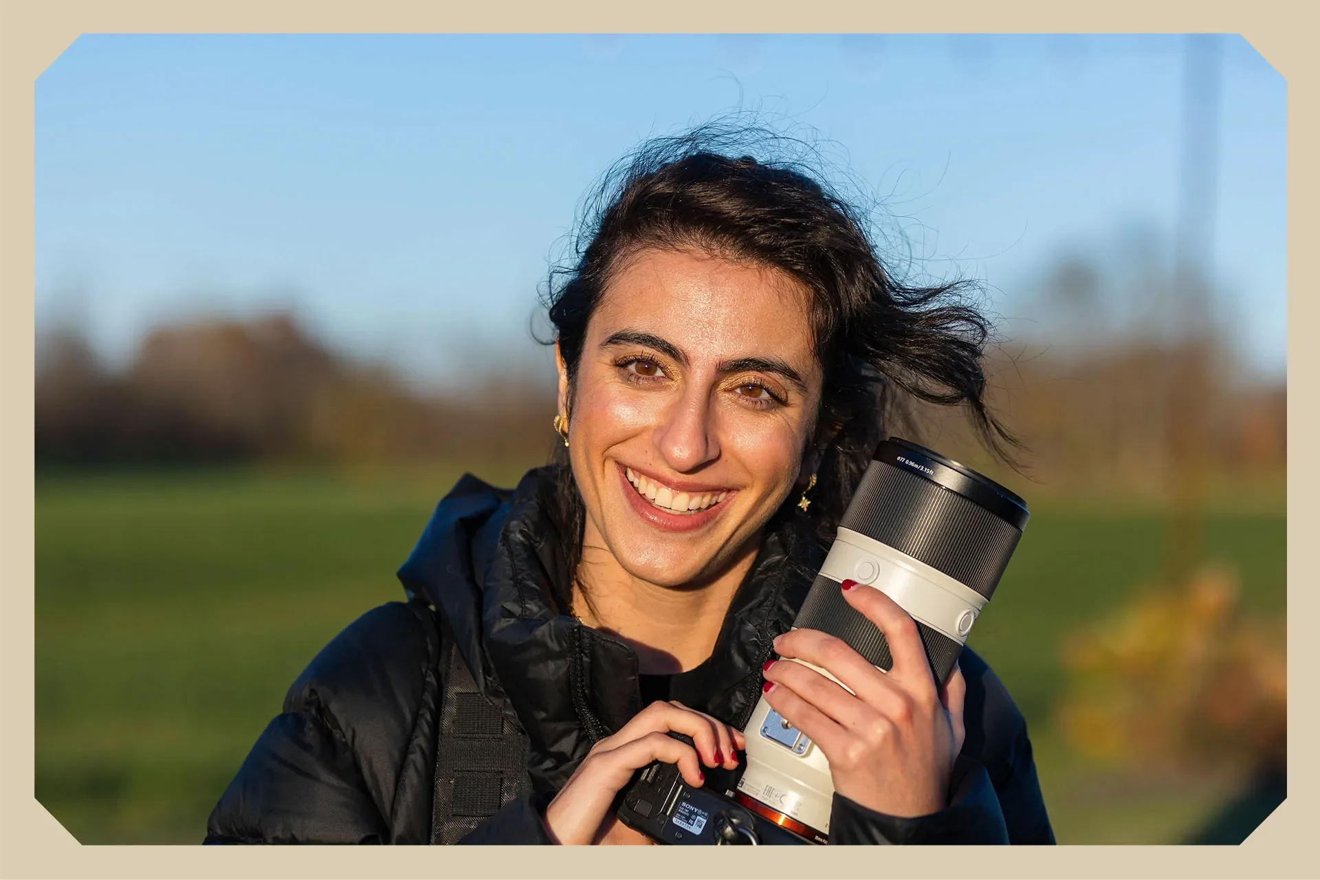 A woman holding a camera with a large zoom lens, smiling outdoors on a sunny day, with a blurred green landscape in the background.