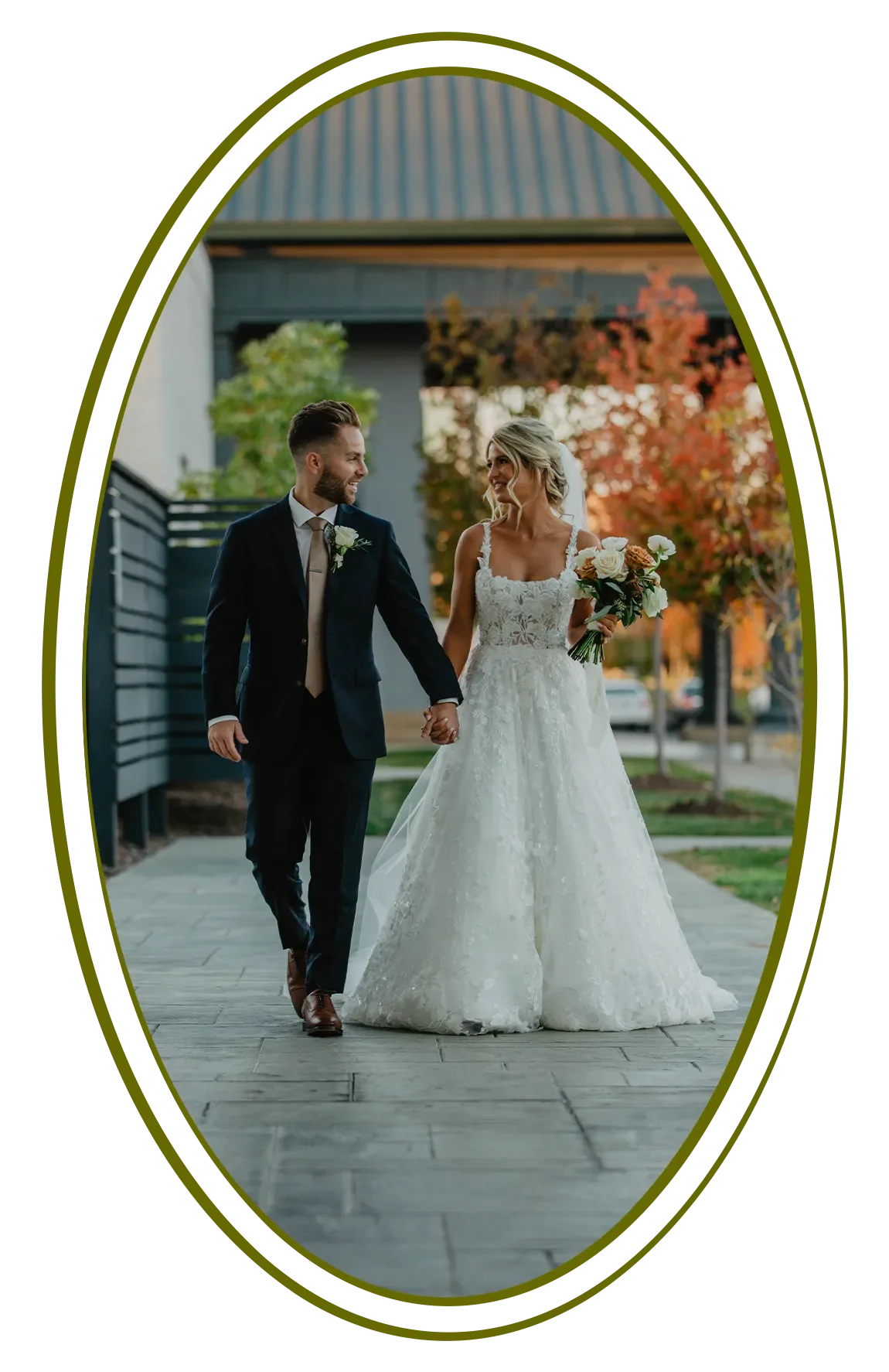 A newlywed couple holding hands and walking outdoors on a walkway with autumn trees in the background. The bride is in a white lace wedding gown holding a bouquet, and the groom is in a dark suit with a boutonniere.