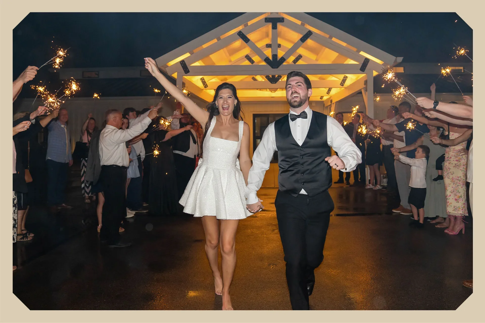 A newlywed couple walks hand-in-hand through a celebration with sparklers held by friends and family at night, illuminated by warm lights on a wooden pavilion.