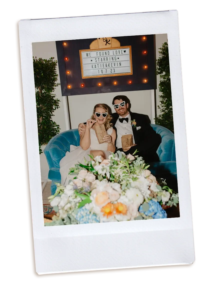 A couple in wedding attire sitting on a blue couch with a large bouquet of flowers in front of them. They are laughing and wearing sunglasses shaped like hearts; the image is in a polaroid frame.