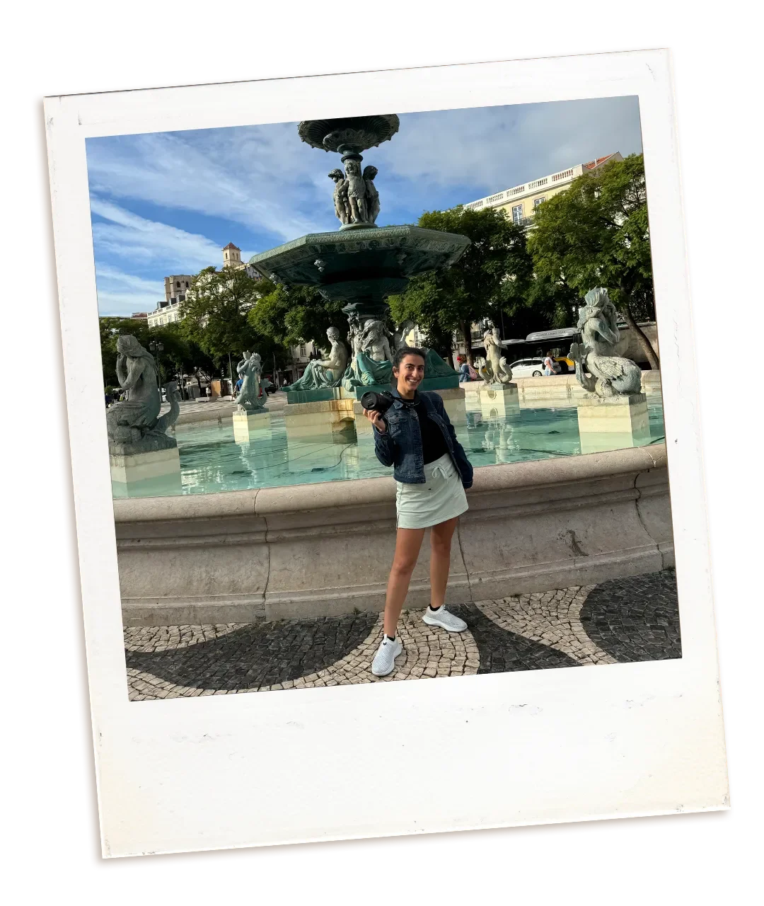 Woman standing in front of a fountain holding a camera, smiling, with trees and buildings in the background.