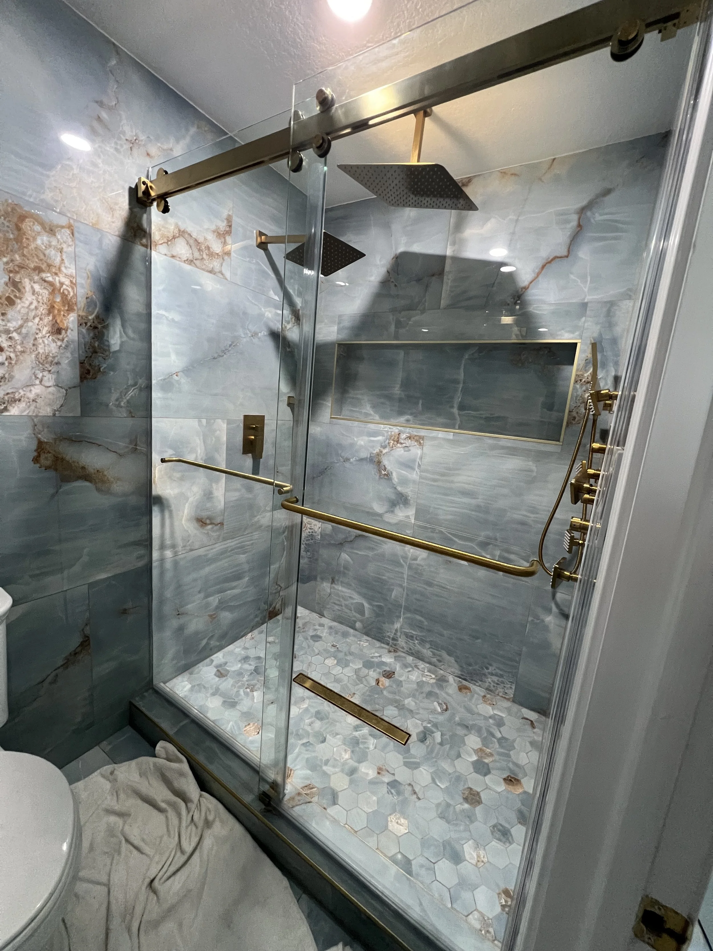 Luxurious walk-in shower with marble tile walls, gold fixtures, and overhead rainfall showerheads, enclosed by glass panels.