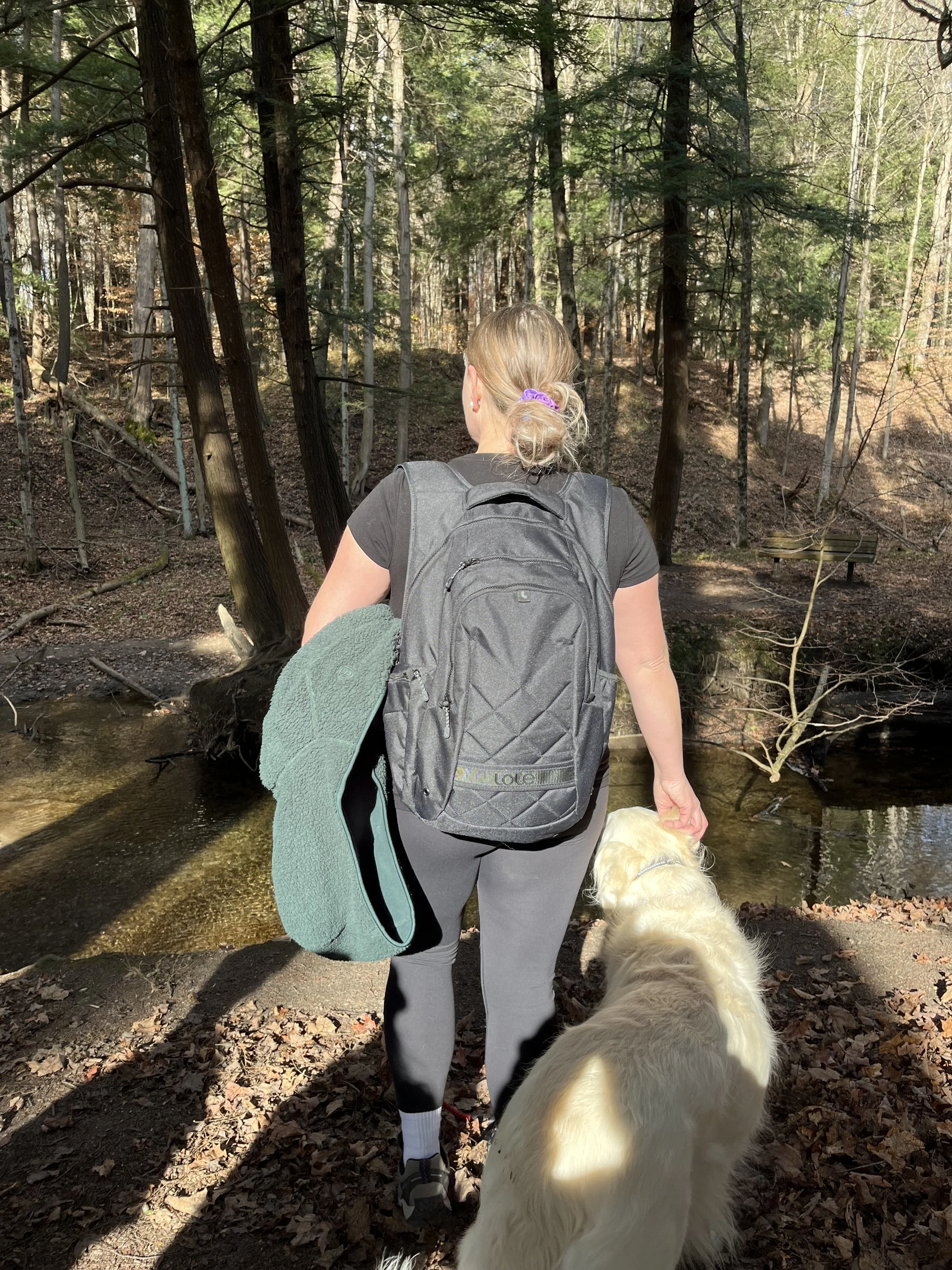 A woman with blonde hair tied in a ponytail, wearing a black t-shirt and gray leggings, holding a leash attached to a golden retriever dog, standing near a stream in a forest during daytime. She carries a gray backpack and a green fleece jacket draped over her arm.
