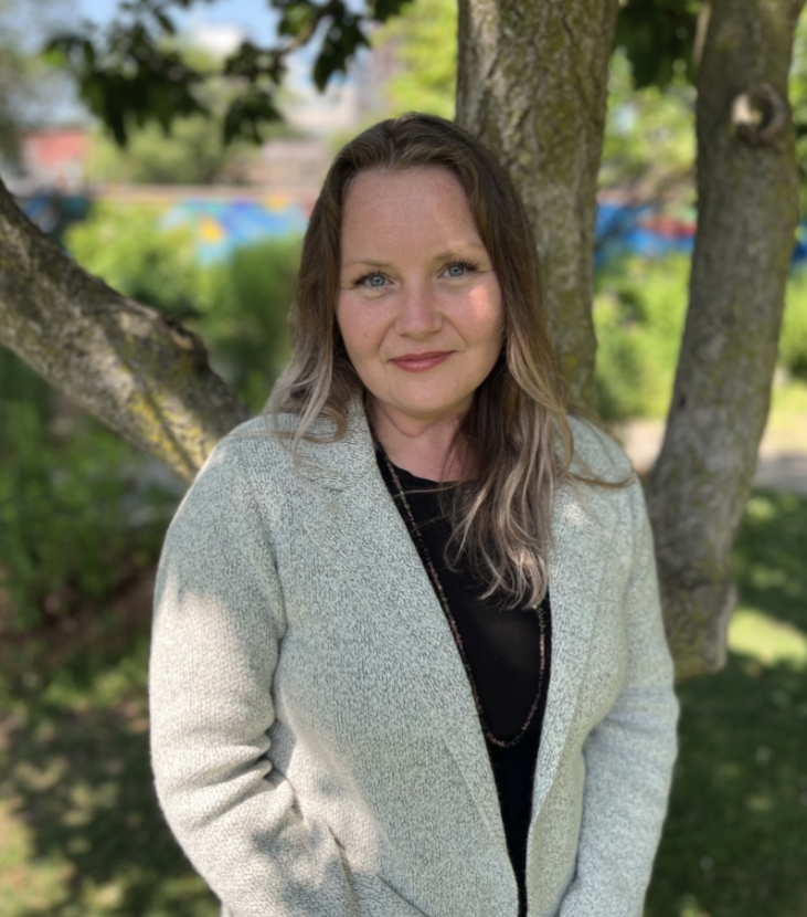 A woman with long, wavy brown hair with blonde highlights, standing outdoors in front of a tree with green leaves. She is wearing a light-colored blazer over a black top and has a necklace.
