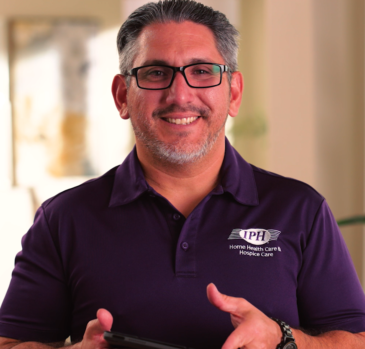A smiling man with glasses and a gray beard, wearing a purple polo shirt with the logo 'IPH Home Health Care & Hospice Care,' holding a tablet, indoors with soft lighting.