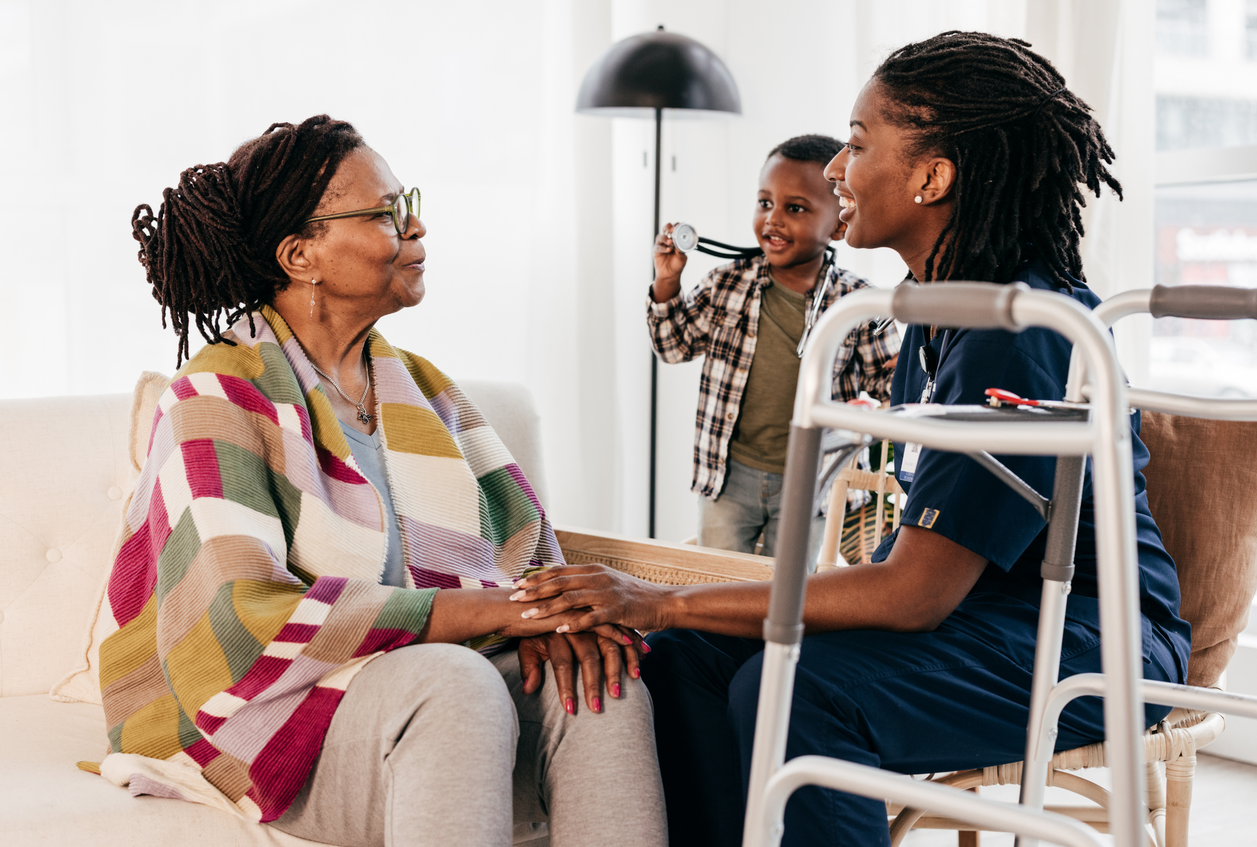 A healthcare professional holding an elderly woman's hand while talking, with a young child in the background holding a stethoscope, inside a bright room.