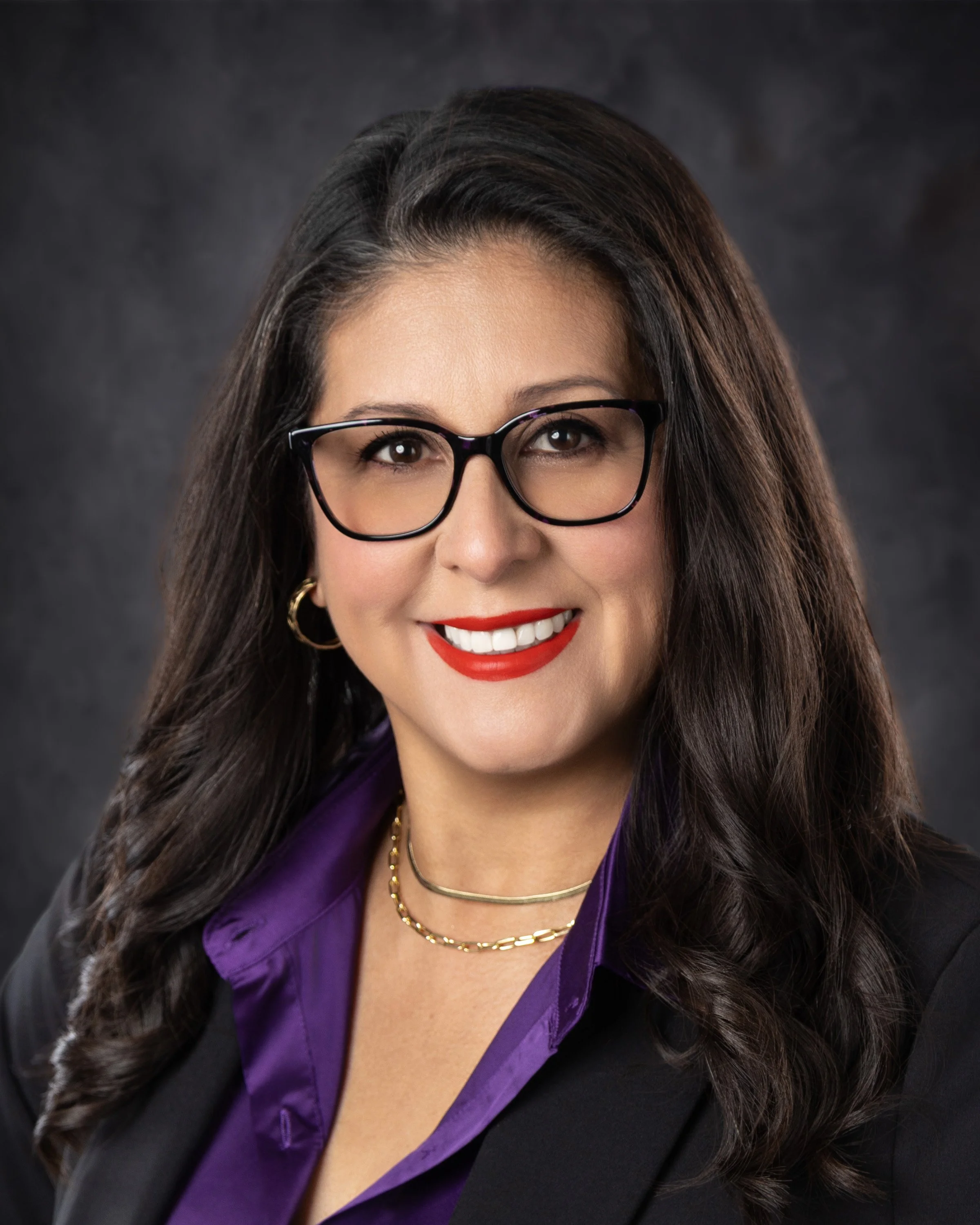 A professional woman smiling, wearing glasses, a purple blouse, black blazer, and gold jewelry against a dark background.