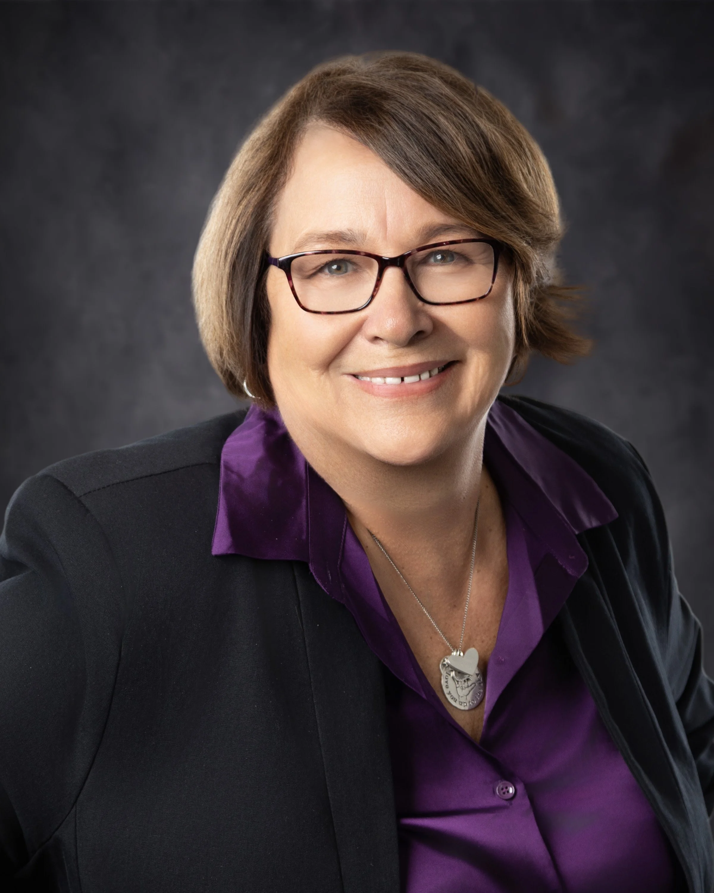 A middle-aged woman with short brown hair, glasses, and fair skin, smiling at the camera. She is wearing a black blazer over a purple shirt with a collar, and a silver necklace with a heart-shaped pendant. The background is dark and blurred.