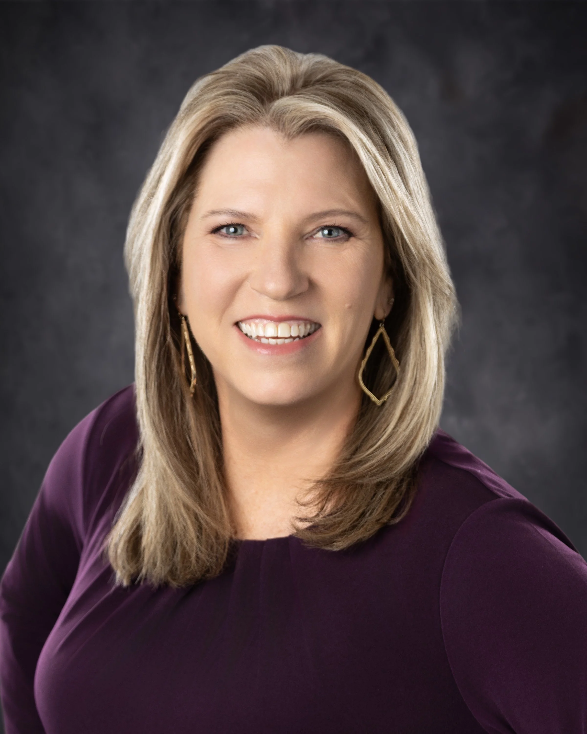 A smiling woman with blonde hair wearing gold hoop earrings and a purple top, posed against a dark background.