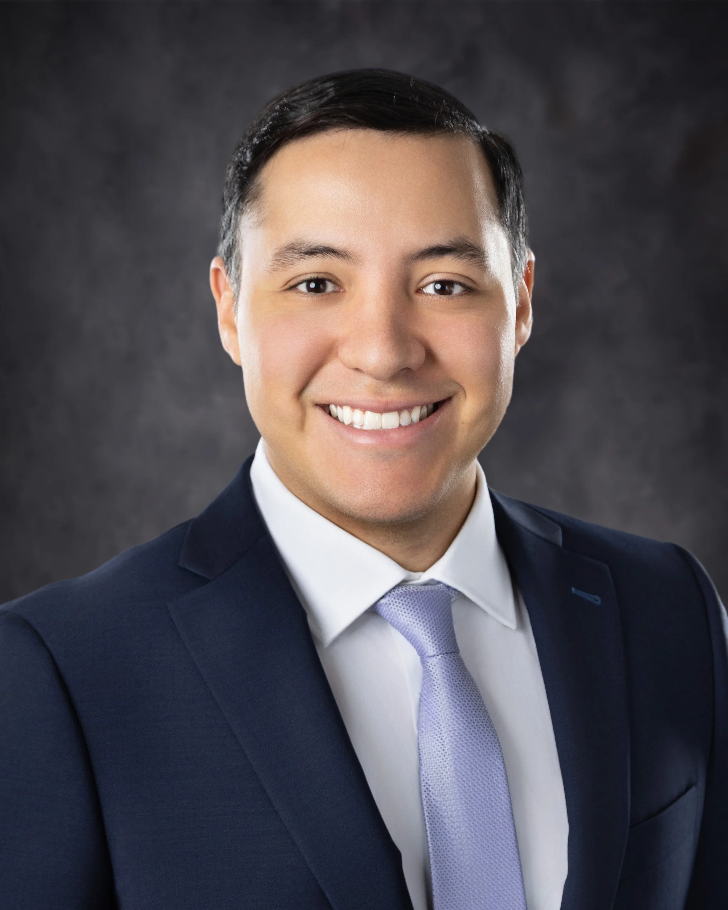 Headshot of a smiling man in a dark suit, light purple tie, and white shirt, against a dark textured background.