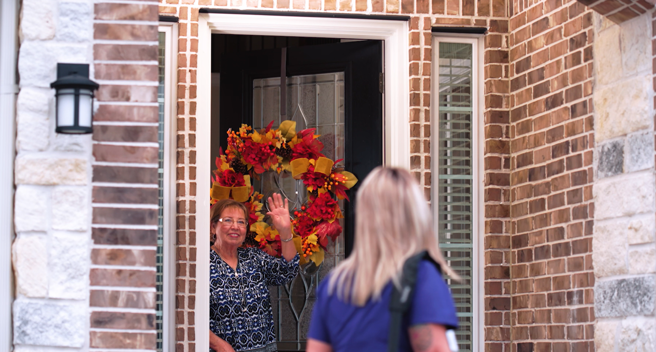 A woman with glasses and a patterned blouse is waving at a person standing outside a brick house with a decorated front door featuring a large fall-themed wreath.