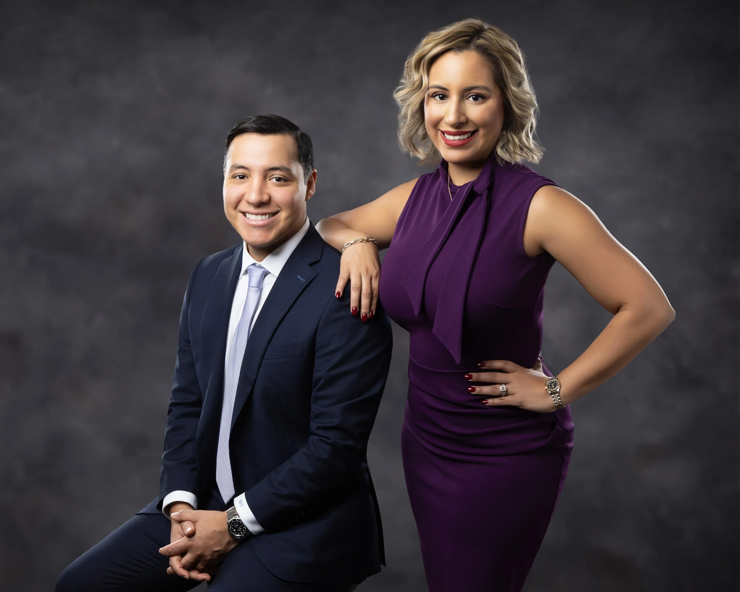 A well-dressed man in a dark suit and light-colored tie, sitting with hands clasped, smiling, and a woman in a sleeveless purple dress standing beside him with her arm resting on his shoulder, smiling, against a dark gray background.