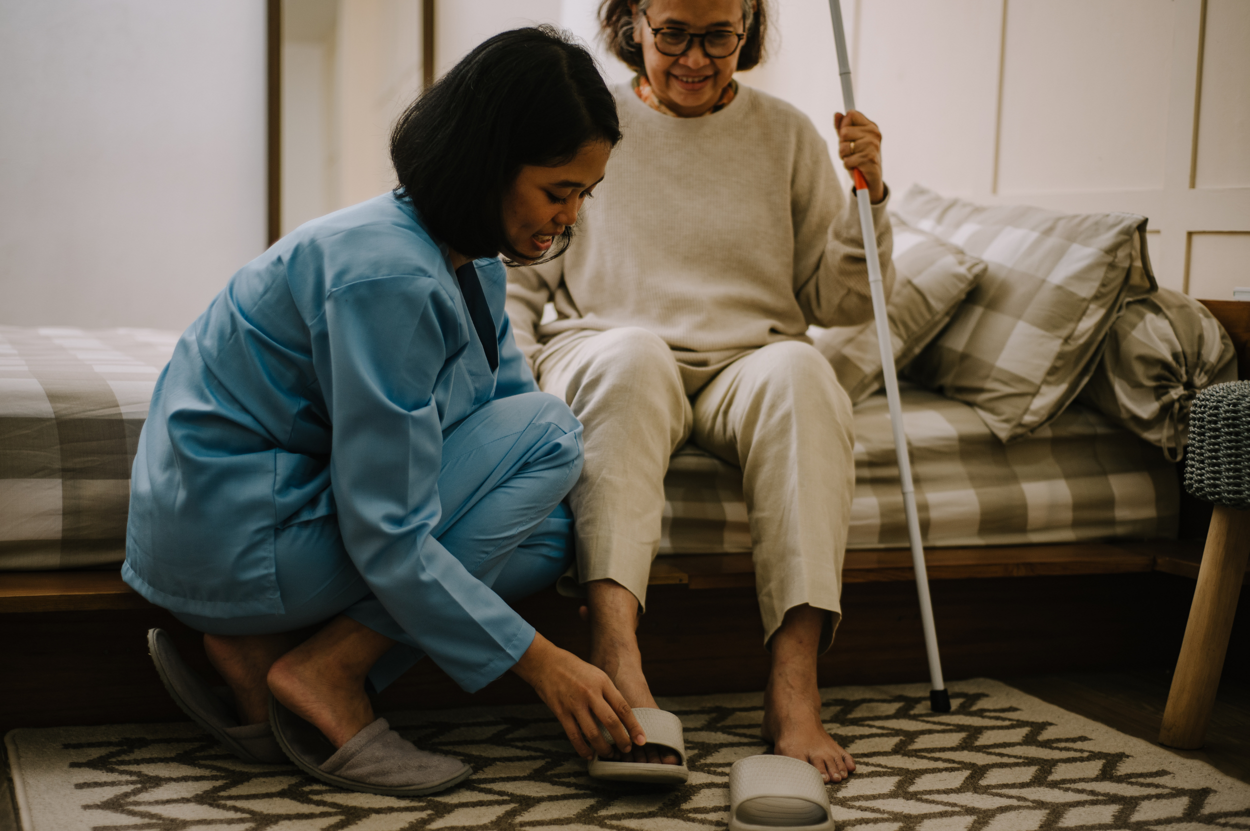 A caregiver assisting an elderly woman with a foot injury by putting on her sandal in a cozy room.