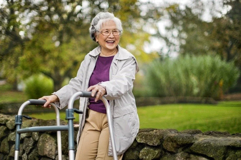 An elderly woman with gray hair and glasses smiling outside while using a walker in a park.