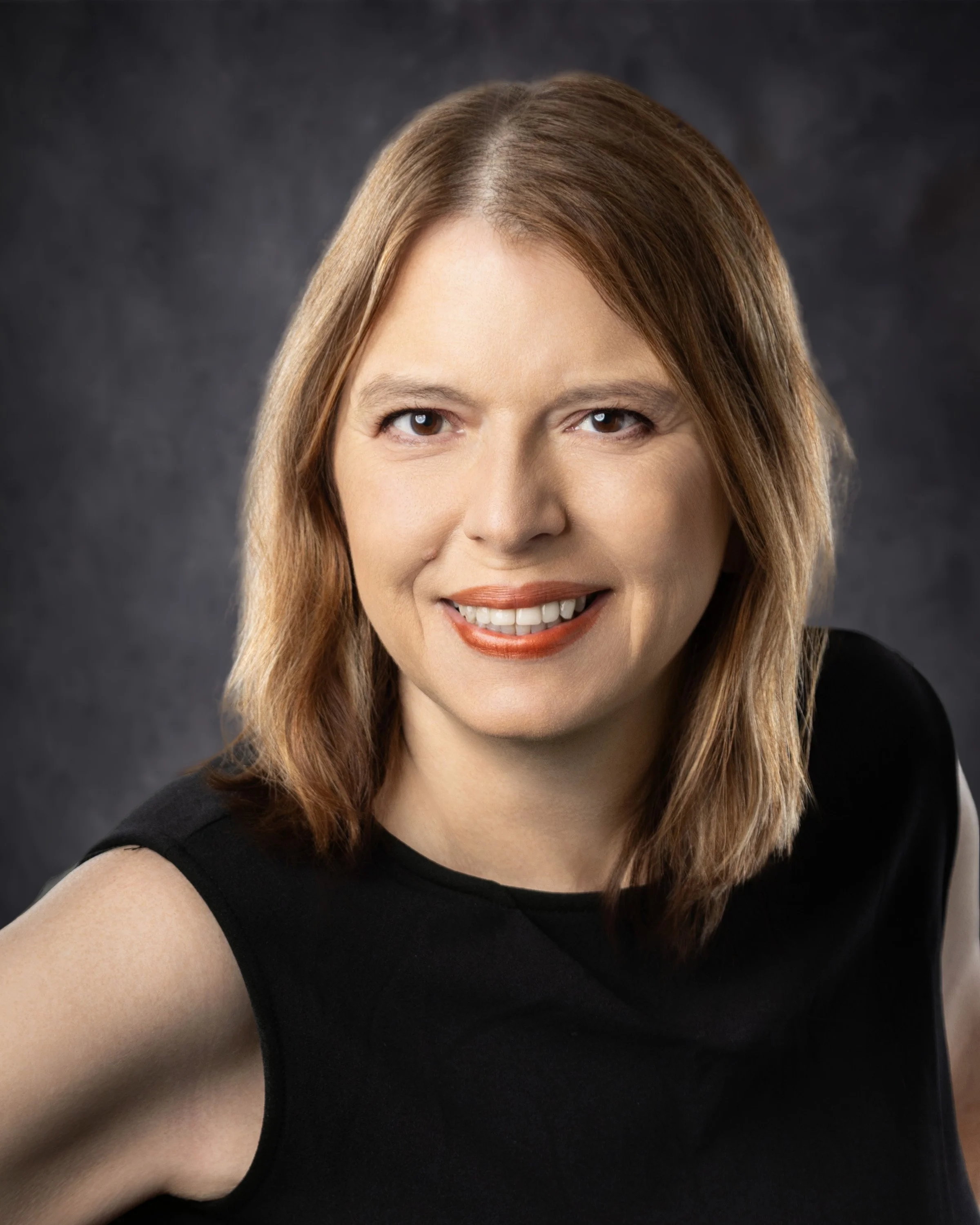 A woman with shoulder-length light brown hair, wearing a black sleeveless top, smiling against a dark gray background.