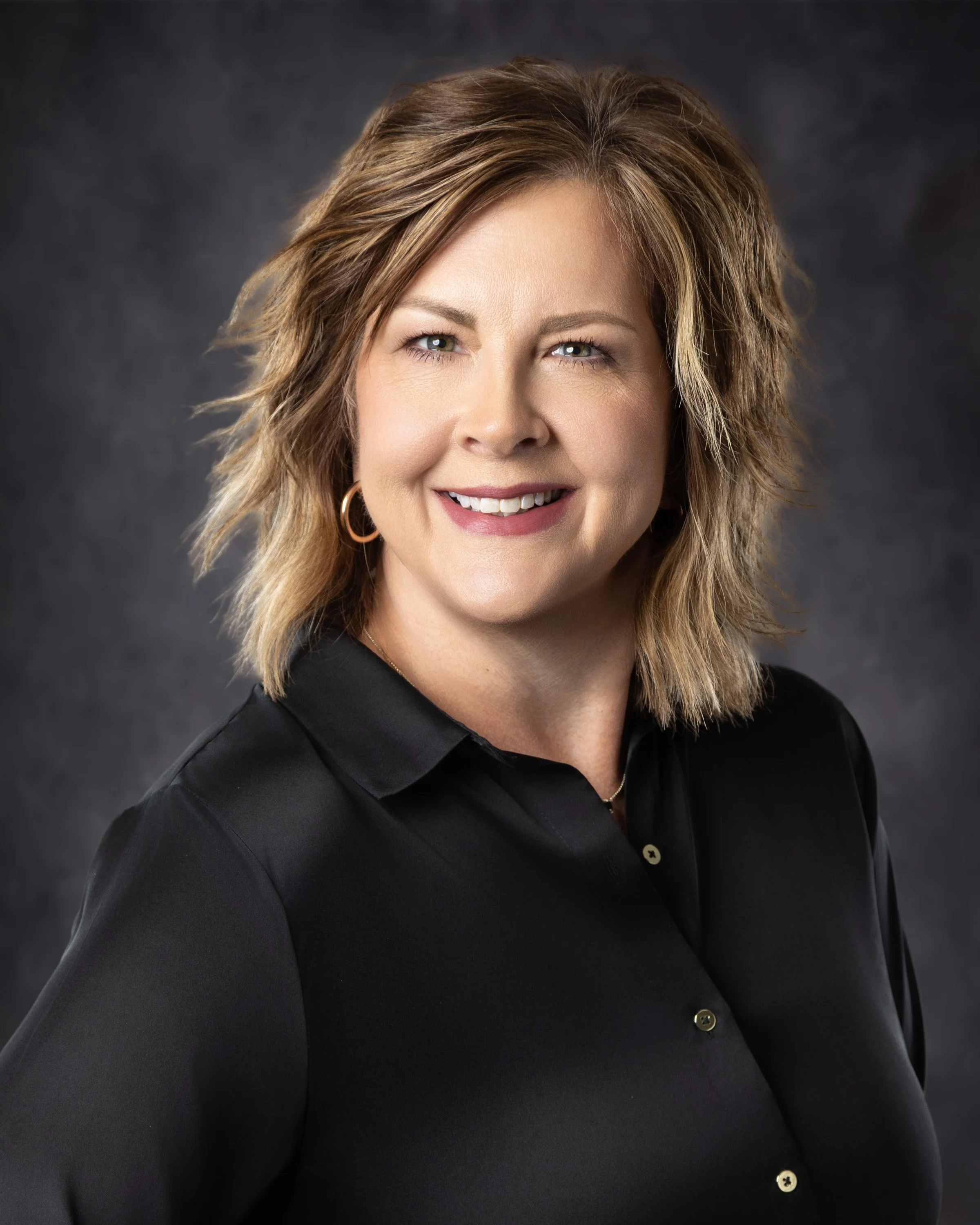 A woman with shoulder-length, wavy light brown hair wearing a black button-up shirt, smiling against a dark, textured background.
