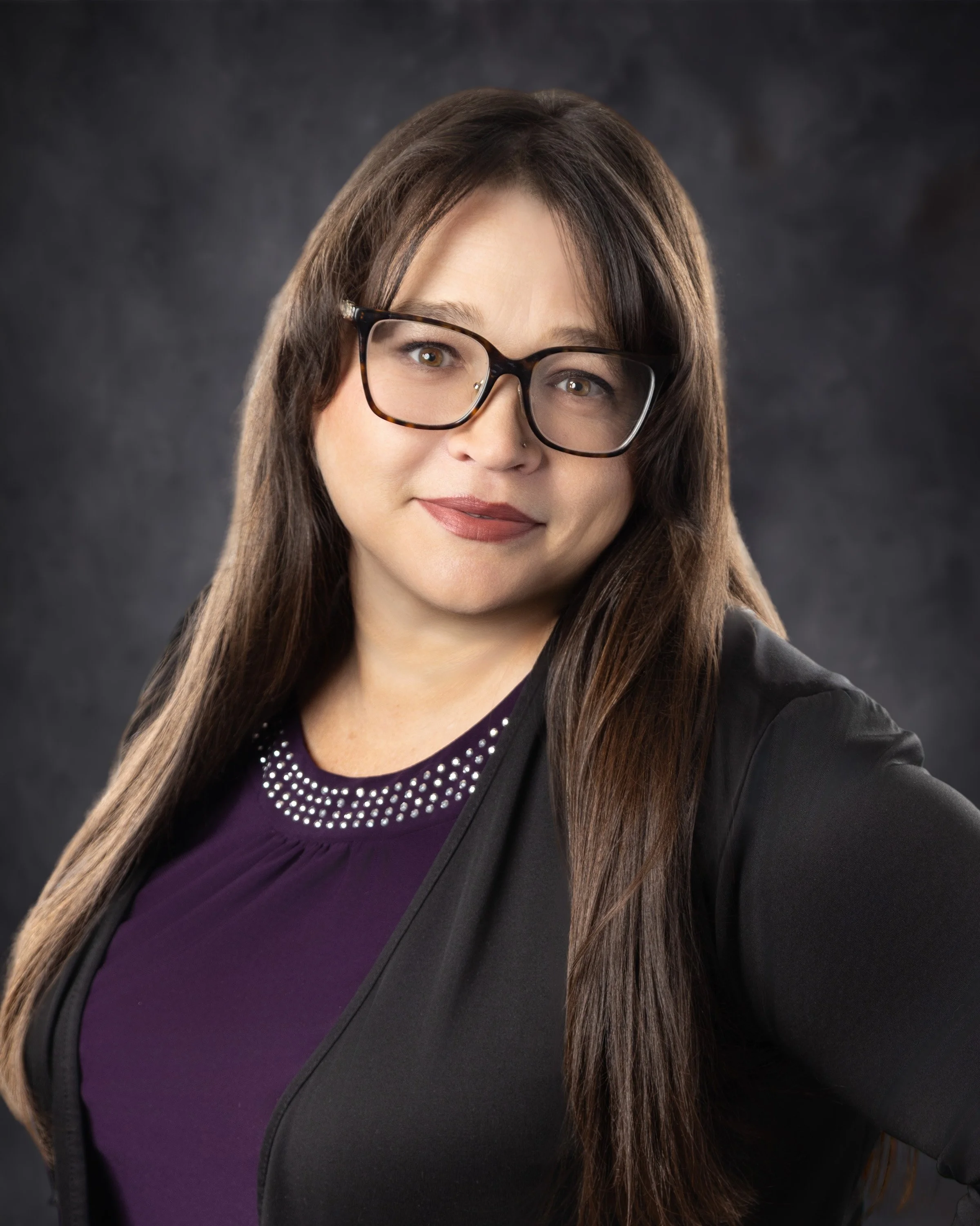 A woman with long brown hair wearing glasses, a purple top with rhinestones on the collar, and a black blazer, smiling against a dark background.