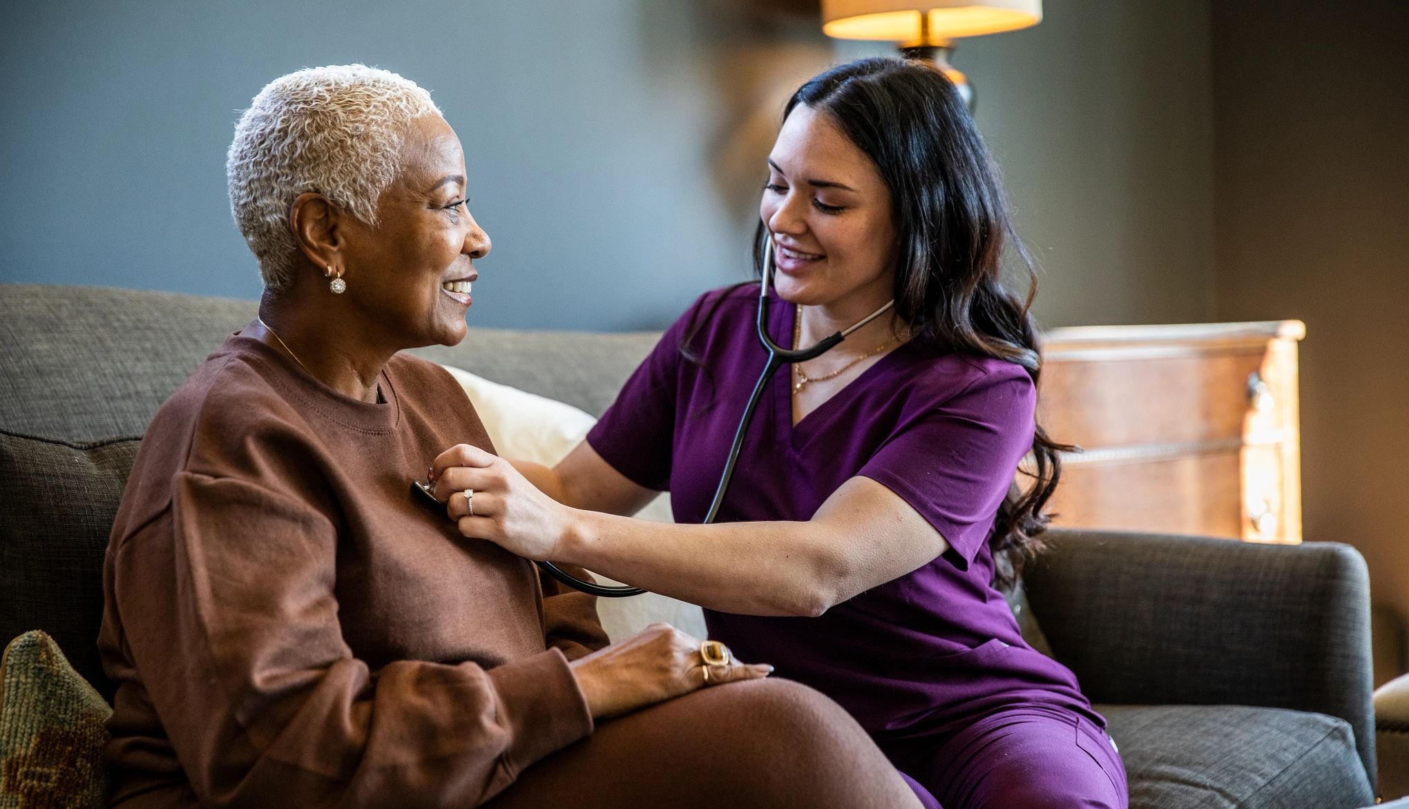 A healthcare professional, wearing purple scrubs and a stethoscope, examines and talks with an elderly woman with short white hair and earrings, sitting on a couch in a cozy room with a lamp and wooden furniture.