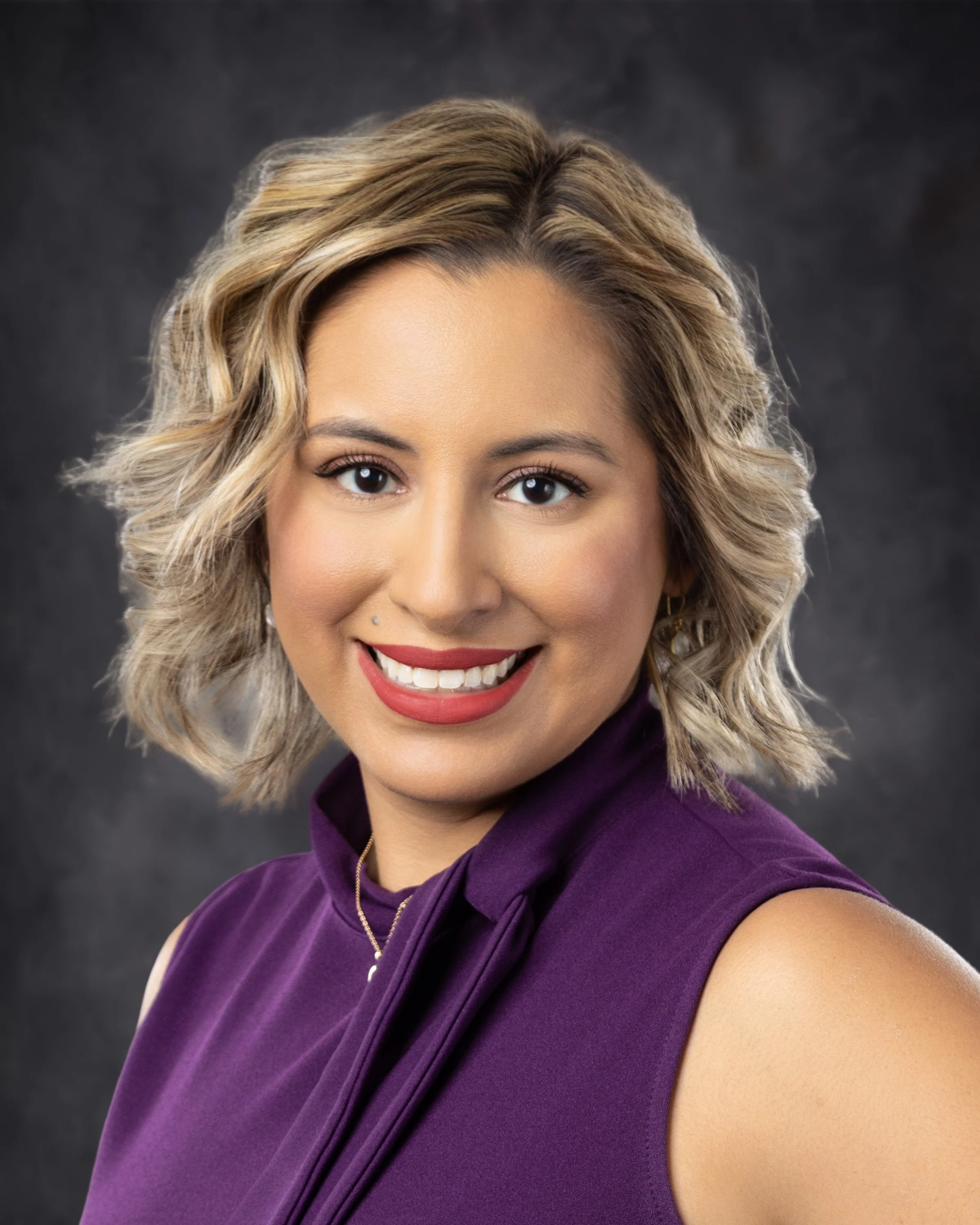 Portrait of a smiling woman with wavy blonde hair wearing a purple sleeveless top, against a dark background.