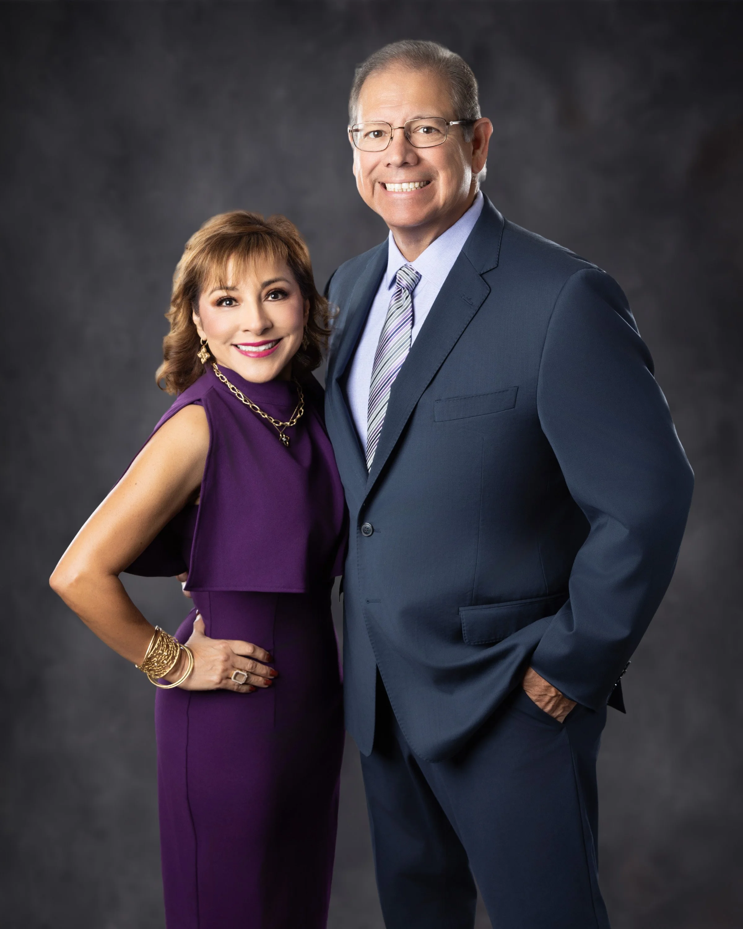 A smiling woman in a purple dress and gold jewelry standing next to a smiling man in a dark suit and tie, both posing against a dark grey background.