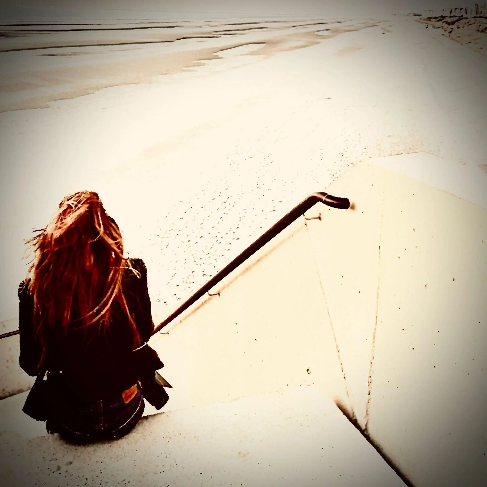 My girl with red hair sitting on stairs at the beach, facing away, with ocean in the background.