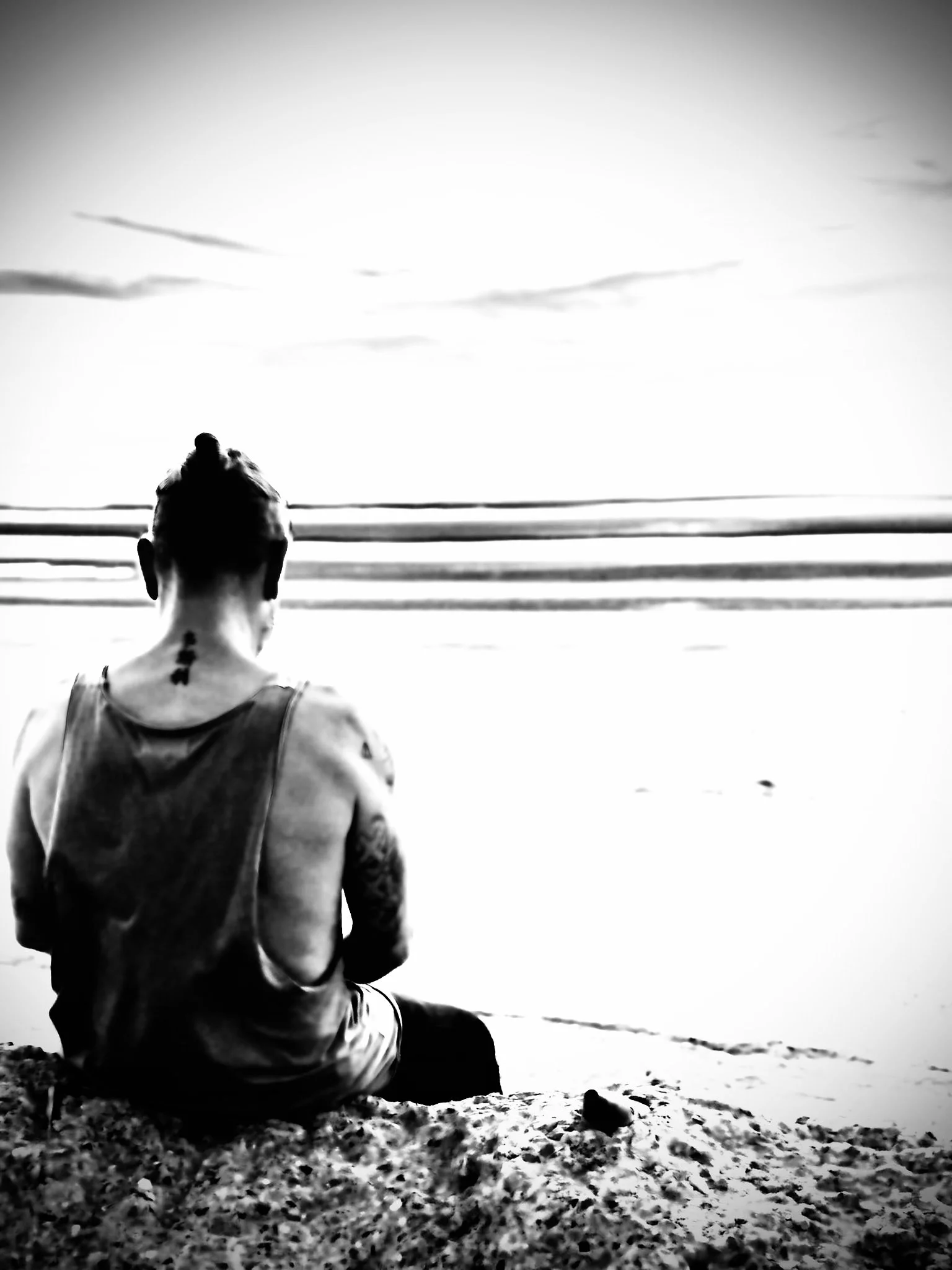 A person with tattoos sits on the sandy beach facing the ocean, with their back to the camera. The photo is in black and white and shows the calm sea with three horizontal lines of waves and a cloudy sky.