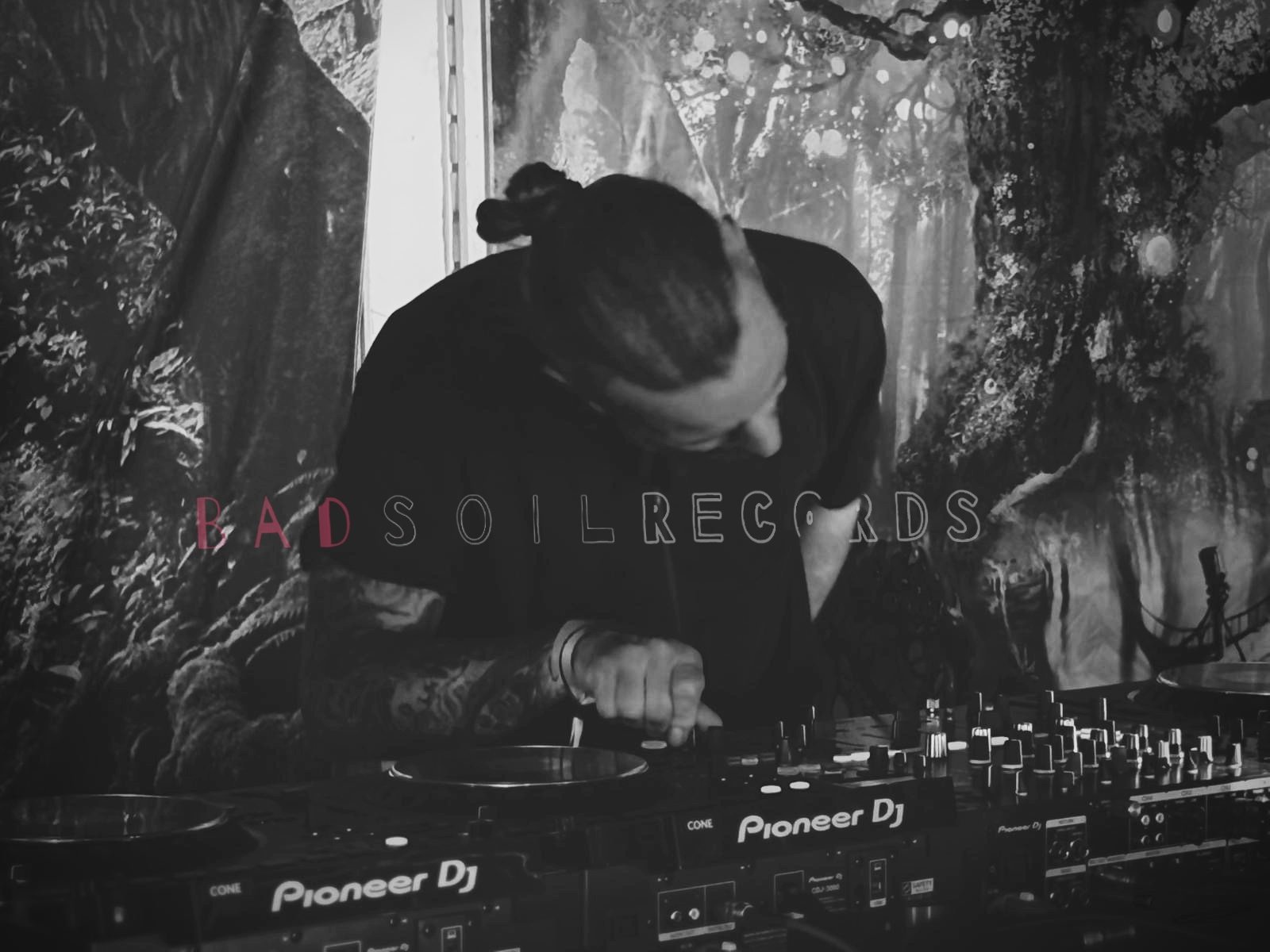 A Belgium based artist called, Wess Vida, label boss with many tattoos and a nomad heart is performing, with Pioneer DJ equipment on a table in front of a crowd in a jungle scene. The words 'BAD SOIL RECORDS' are reflecting his soul into a label.