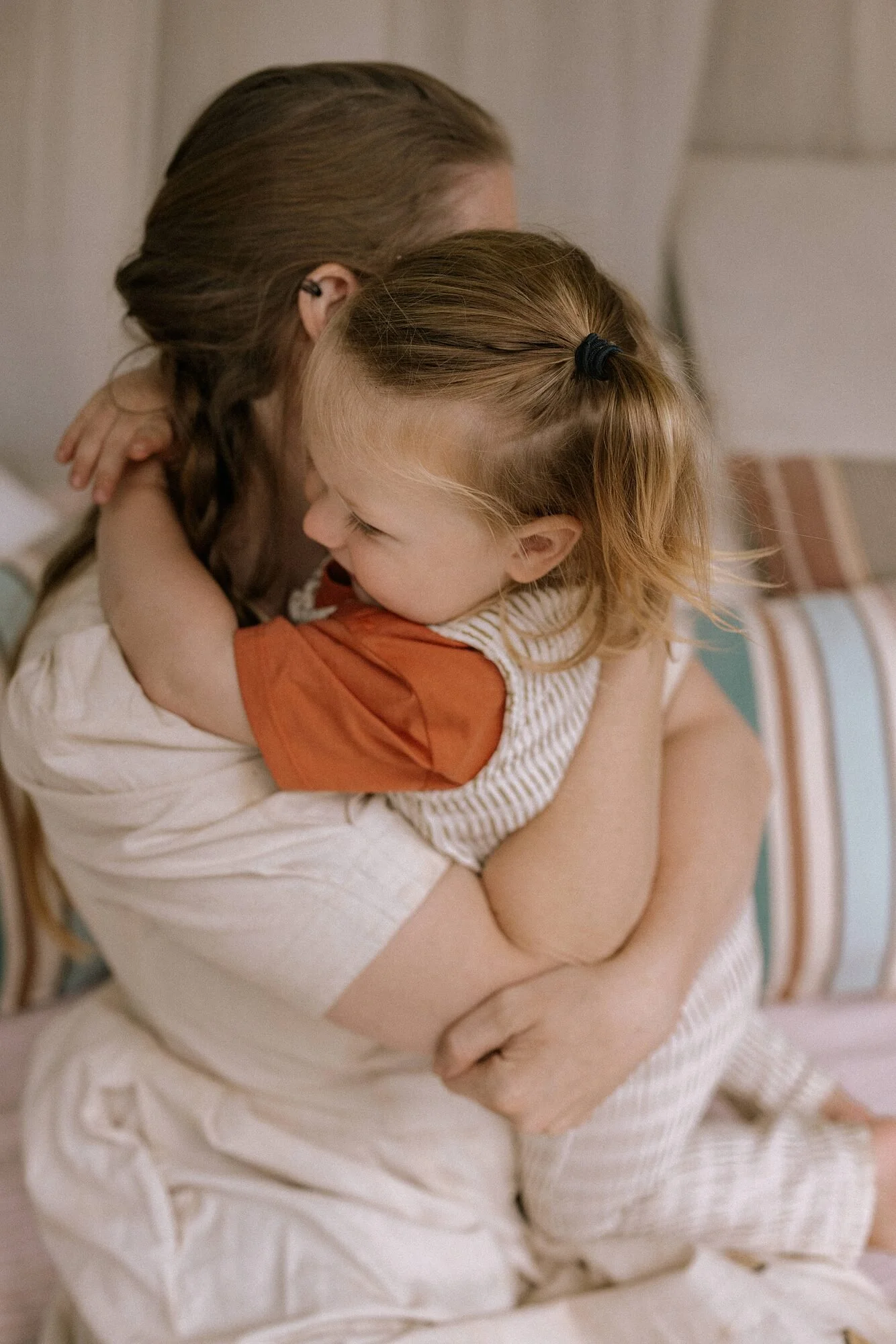 Een persoon met bruin haar en een kind met blond haar in een omhelzing, zittend op een bed in een slaapkamer.