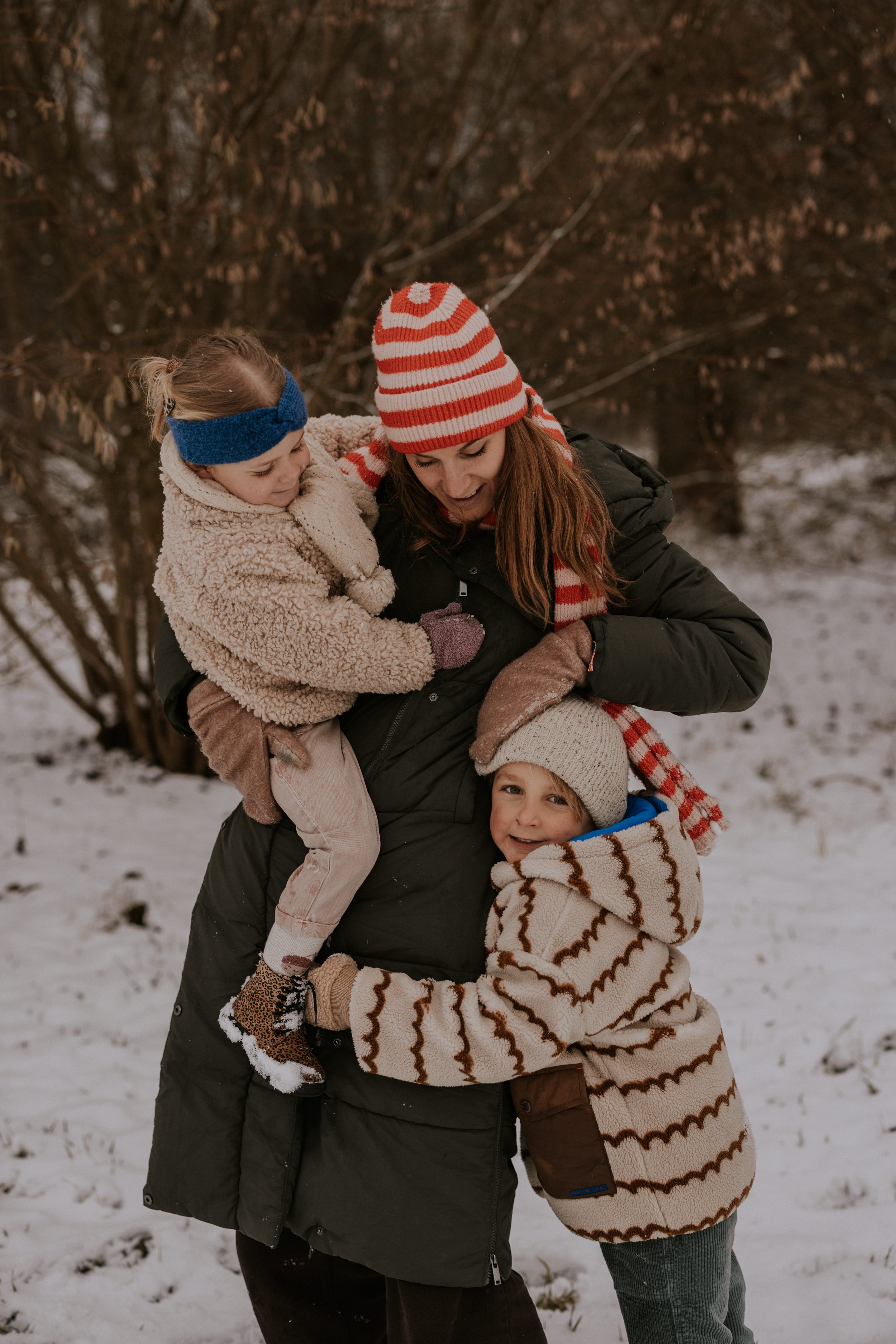 Vrouw met twee jonge kinderen in winterkleding die samen lachen en knuffelen buiten in de sneeuw.