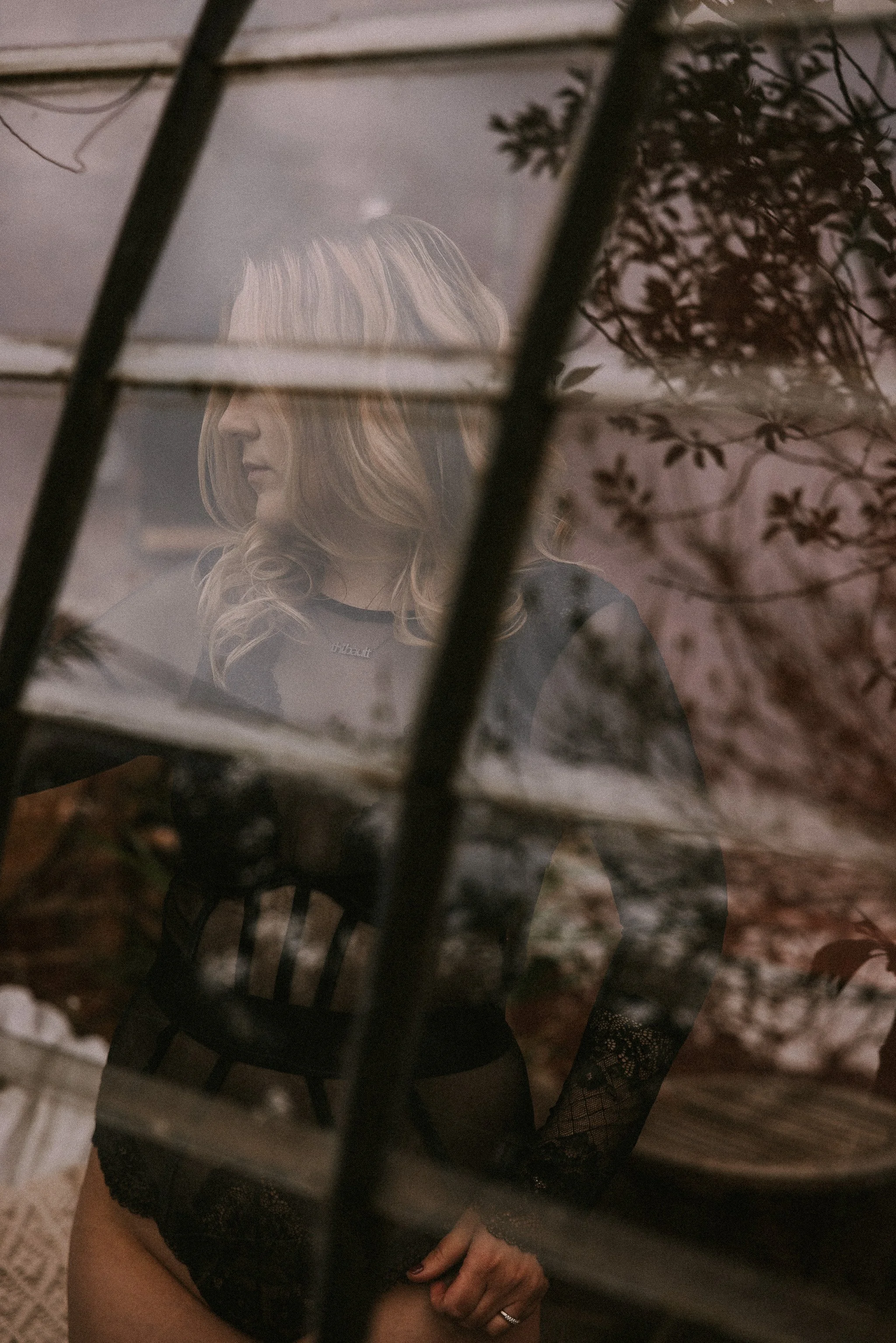 Een vrouw gekleed in zwarte kanten kleding, gezien via een spiegel of raam met horizontale lamellen, met bloemen en planten op de achtergrond.