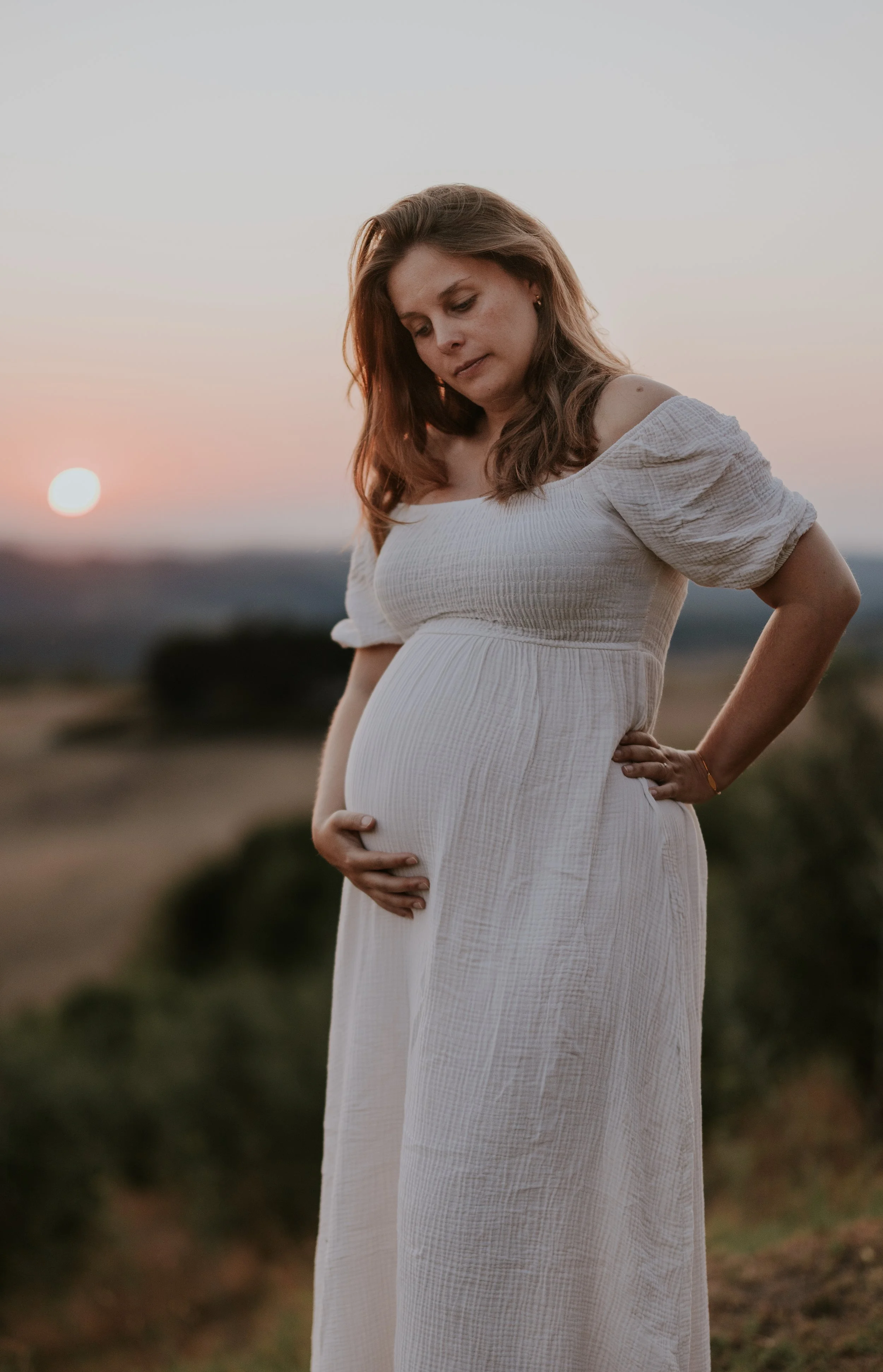 Zwangere vrouw in witte jurk die haar buik vasthoudt tijdens zonsondergang in een open veld.