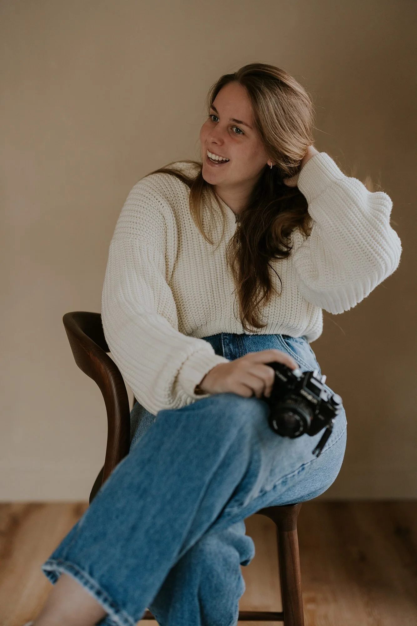 Een glimlachende vrouw met lang bruin haar, die zit op een houten stoel, een camera in haar hand houdt, en gekleed is in een witte gebreide trui en blauwe jeans.
