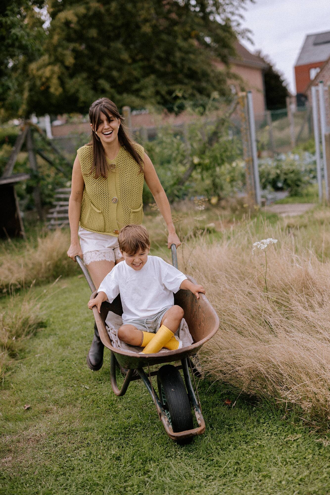 Een vrouw met bruin haar die lachend een kind in een kruiwagen duwt. Het kind zit in de kruiwagen met geel regenlaarzen en witte kleren. Ze zijn buiten op een grasveld met bomen en huizen op de achtergrond.