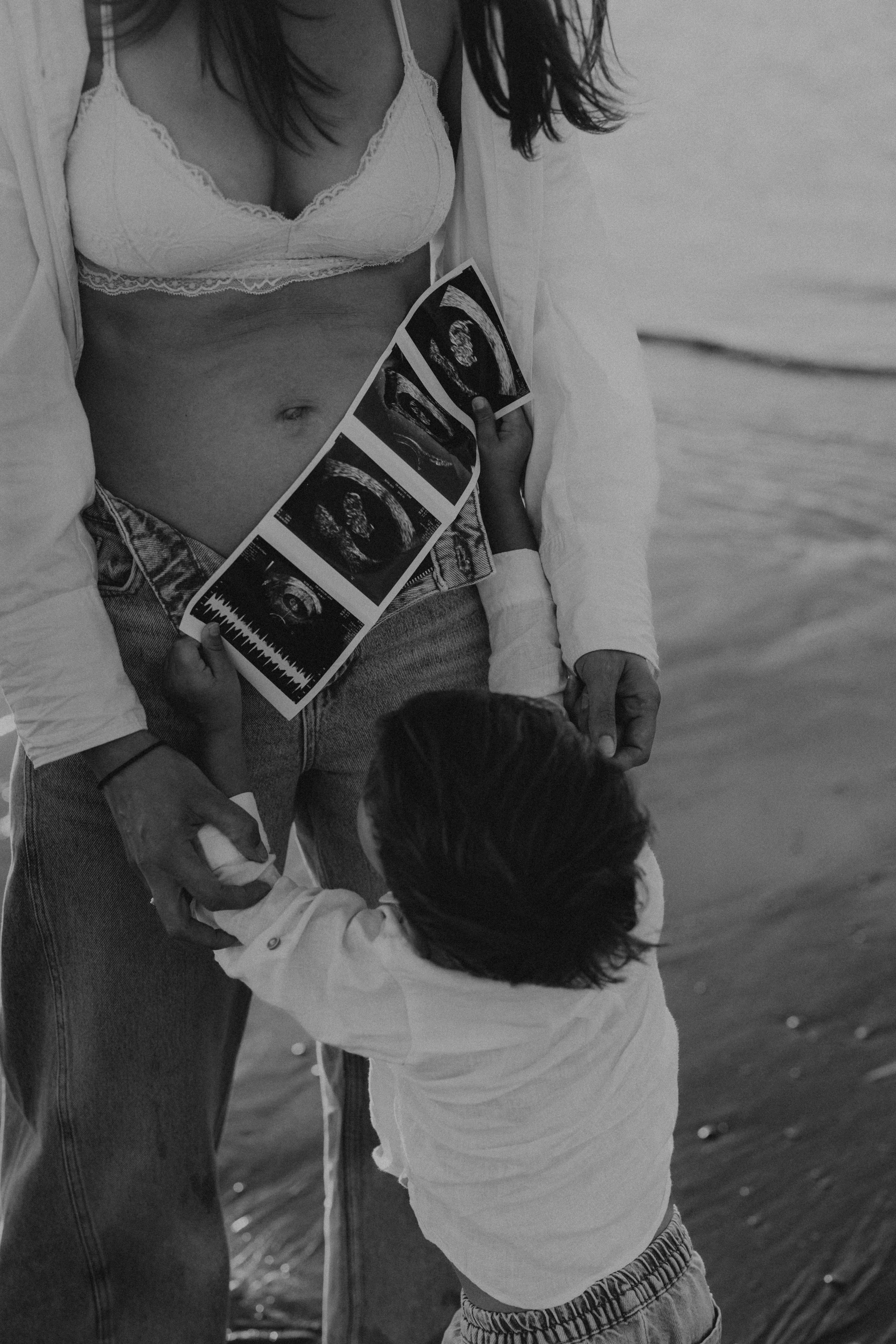 Een zwangere vrouw op het strand houdt een kinder hand vasthouden, terwijl ze haar eerste echo's toont.