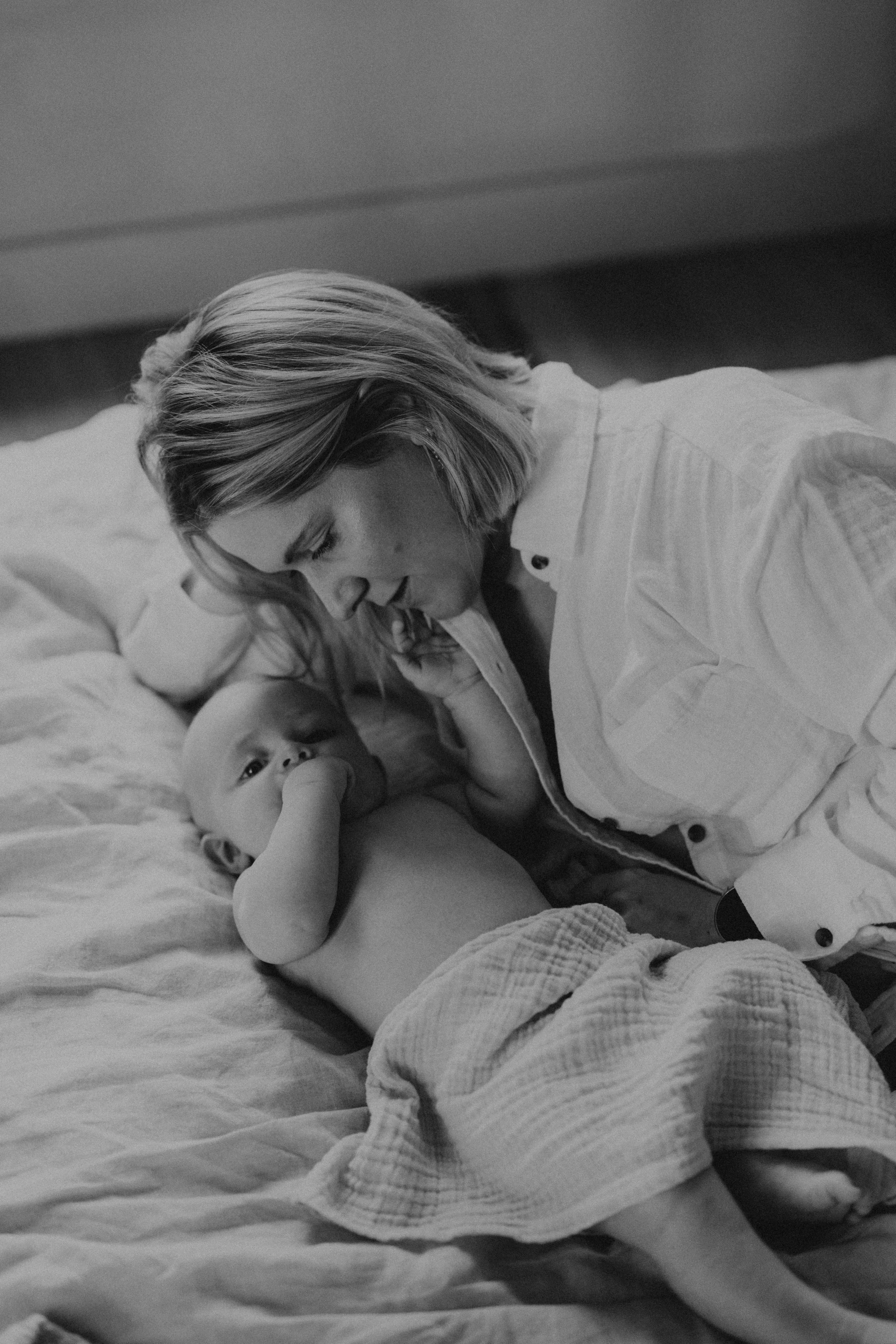 Een vrouw ligt op een bed naast een baby die op de rug ligt, een hand in zijn mond heeft en naar de vrouw kijkt.