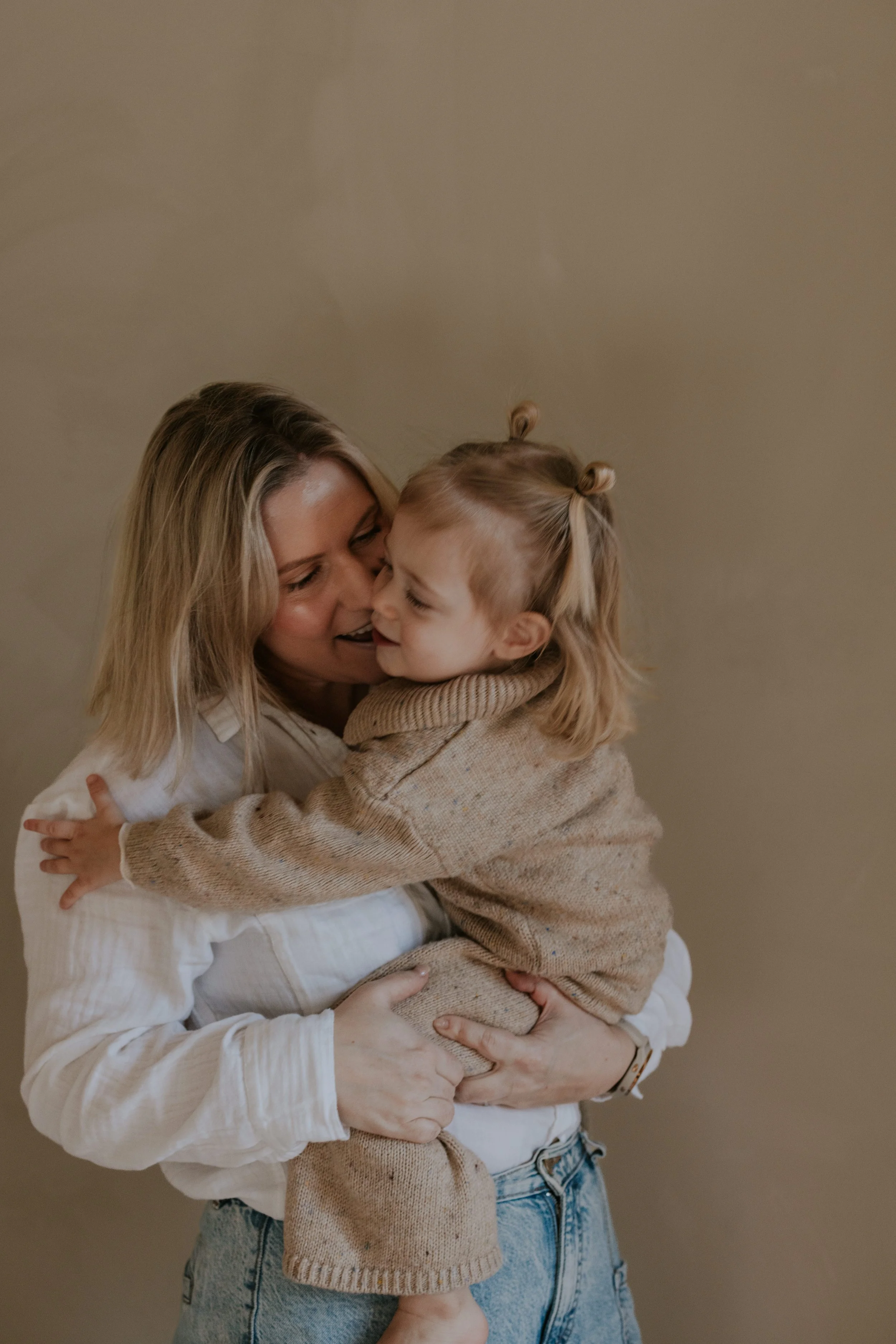 Vrouw houdt lachend een klein meisje in haar armen, beiden glimlachen en knuffelen elkaar, binnen tegen een effen beige achtergrond.