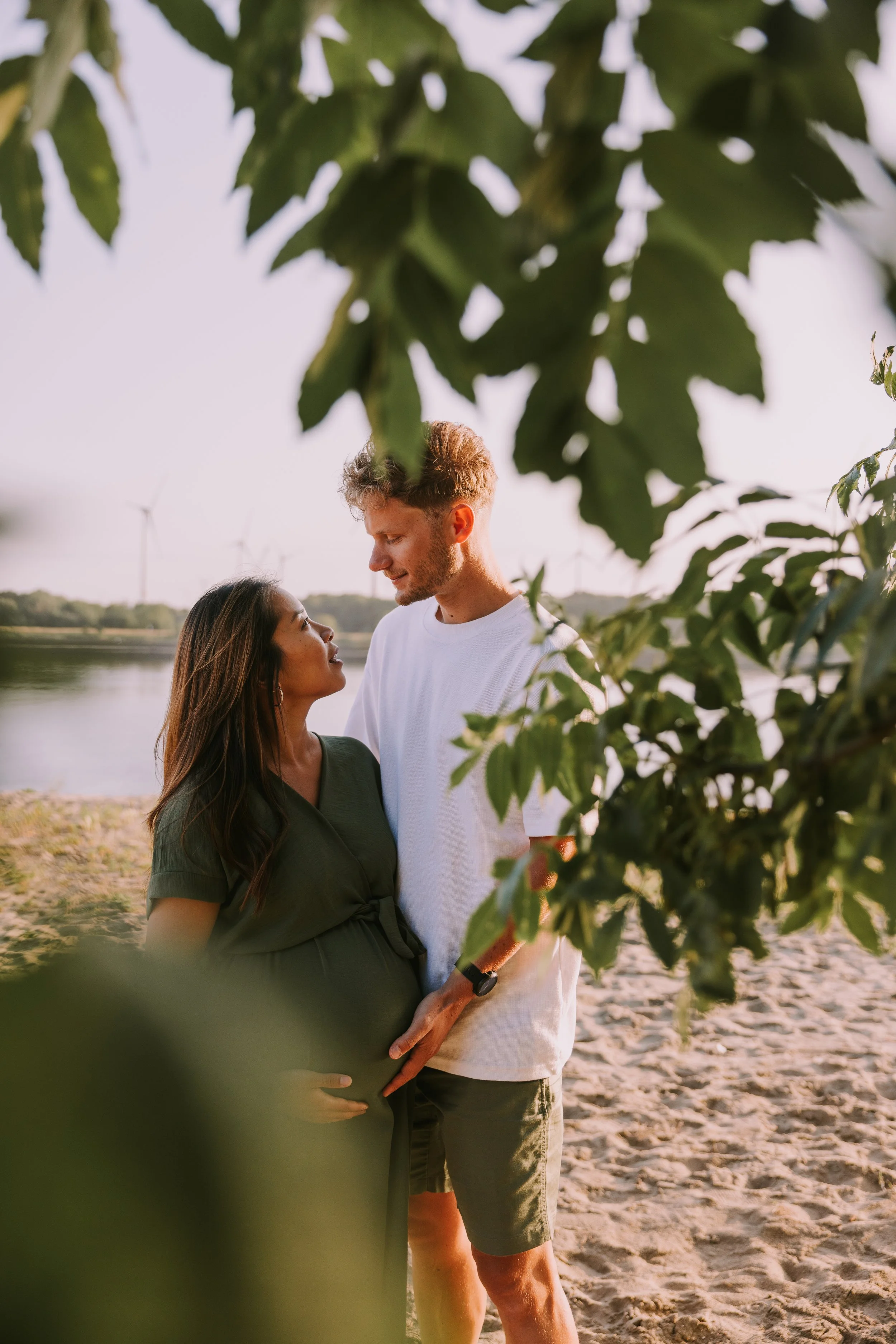 Een zwangere vrouw en haar partner staan dicht bij elkaar op een strand met water en windmolens op de achtergrond, omgeven door groene bladeren.