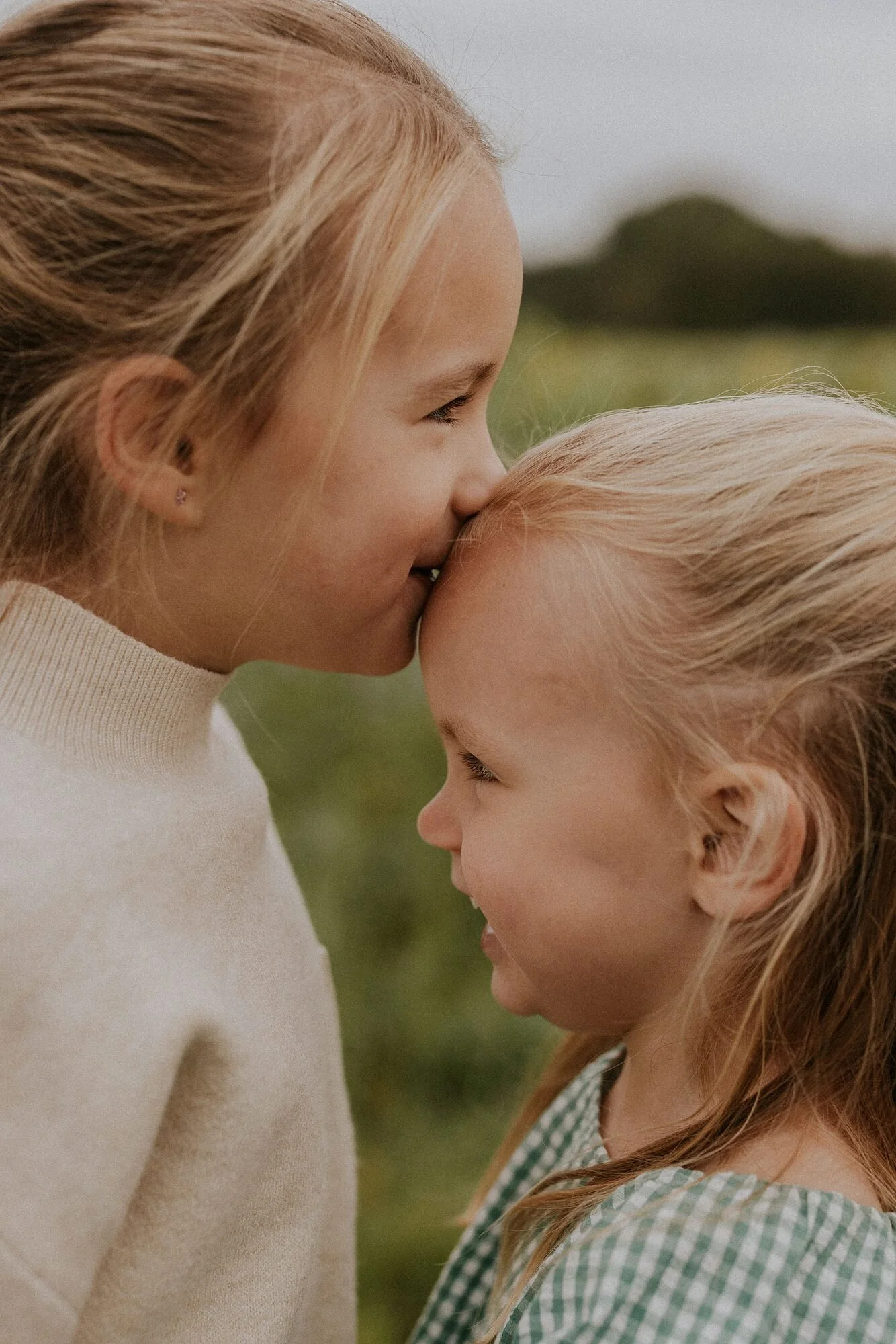 Een jonge vrouw en een jong meisje drukken hun voorhoofden tegen elkaar en glimlachen in een groene buitenomgeving.