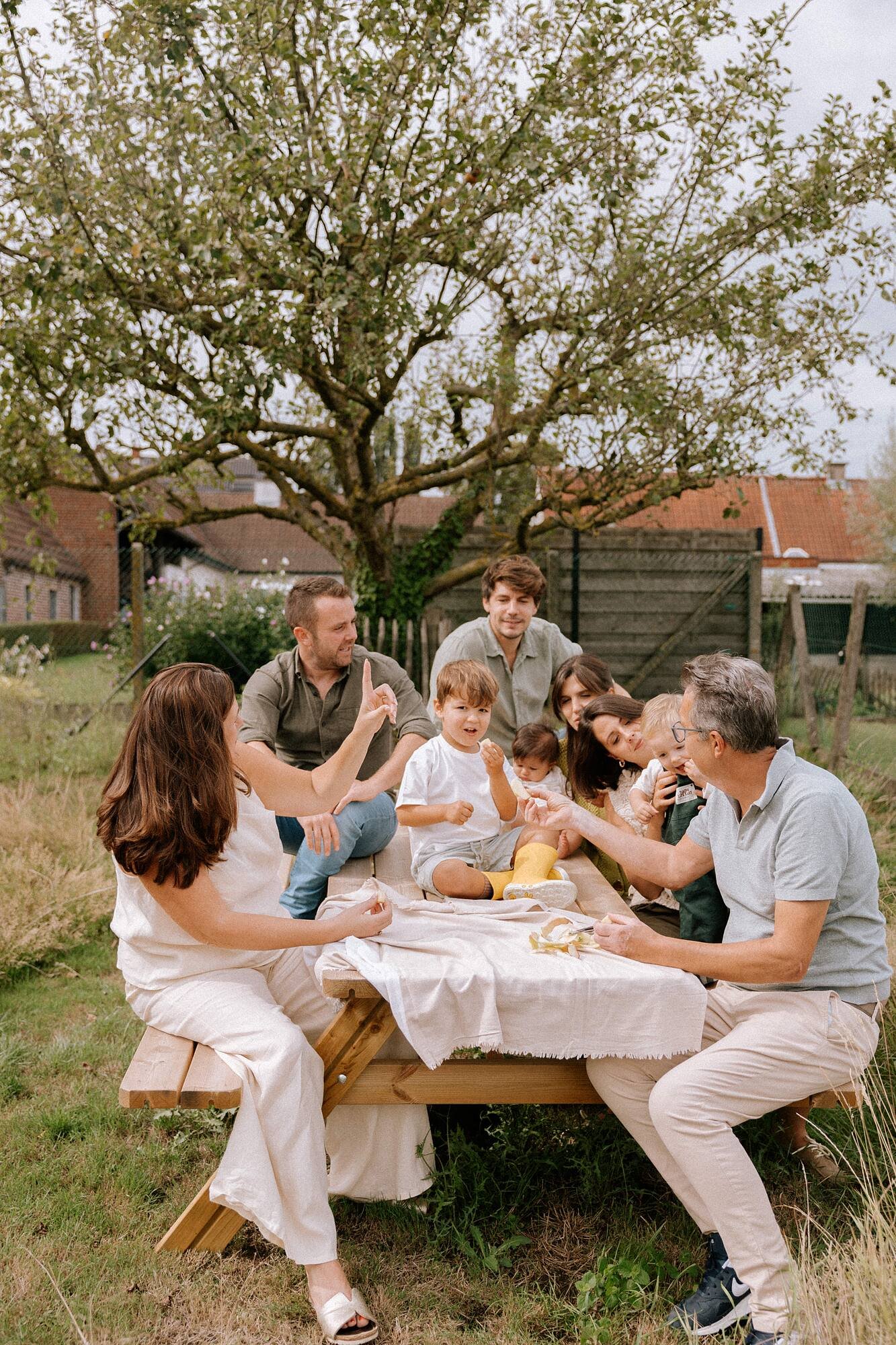 Gezinnen en kinderen genieten samen van een buitenmaaltijd onder een grote boom in een tuin. Ze zitten aan een lange houten tafel met een witte tafelkleed, omgeven door gras en een schutting. Het lijkt een gezellige zomerse middag.