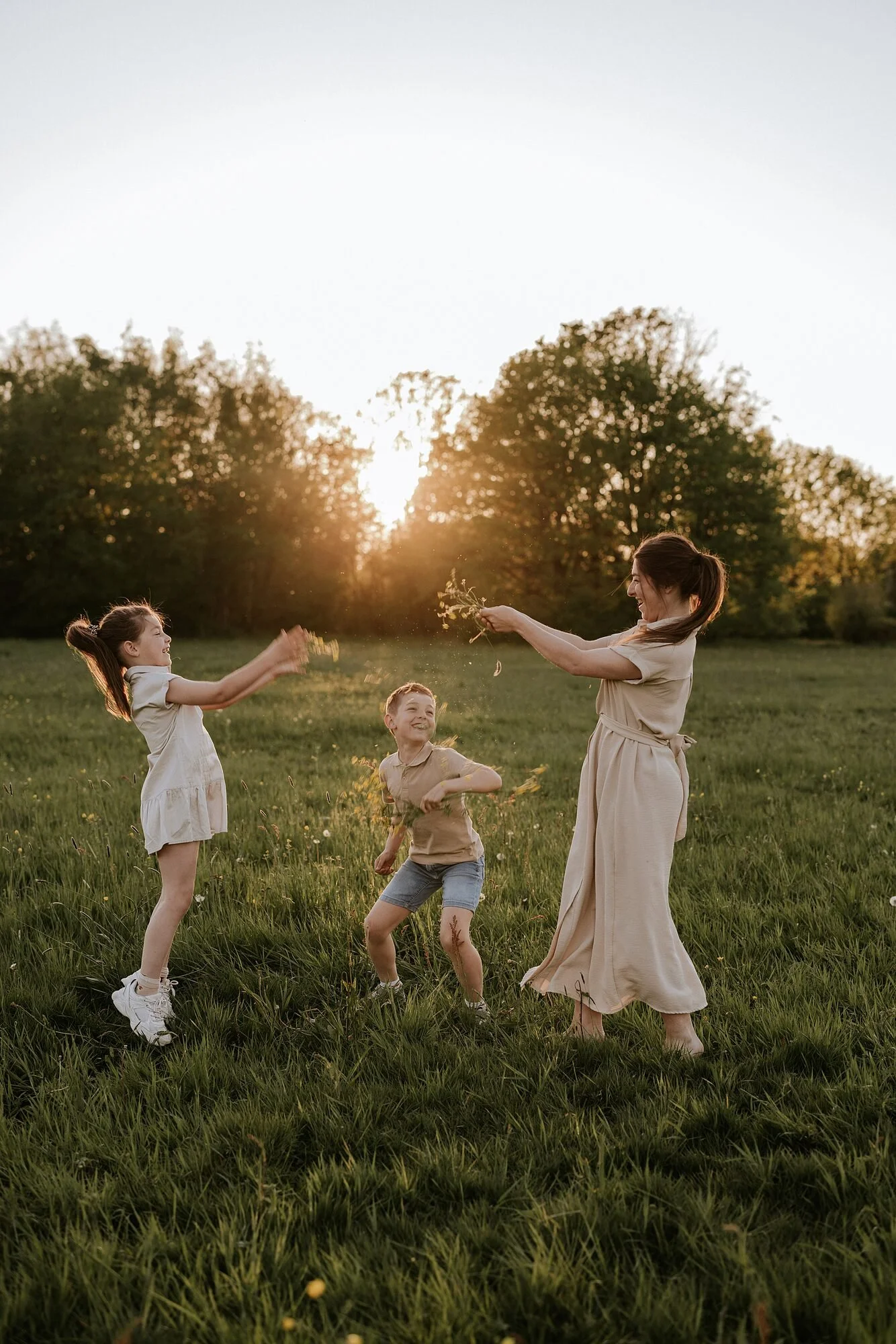 Vrouw en kinderen spelen en lachen in een open grasveld bij zonsondergang, met bomen op de achtergrond, terwijl ze met water en gras spelen.