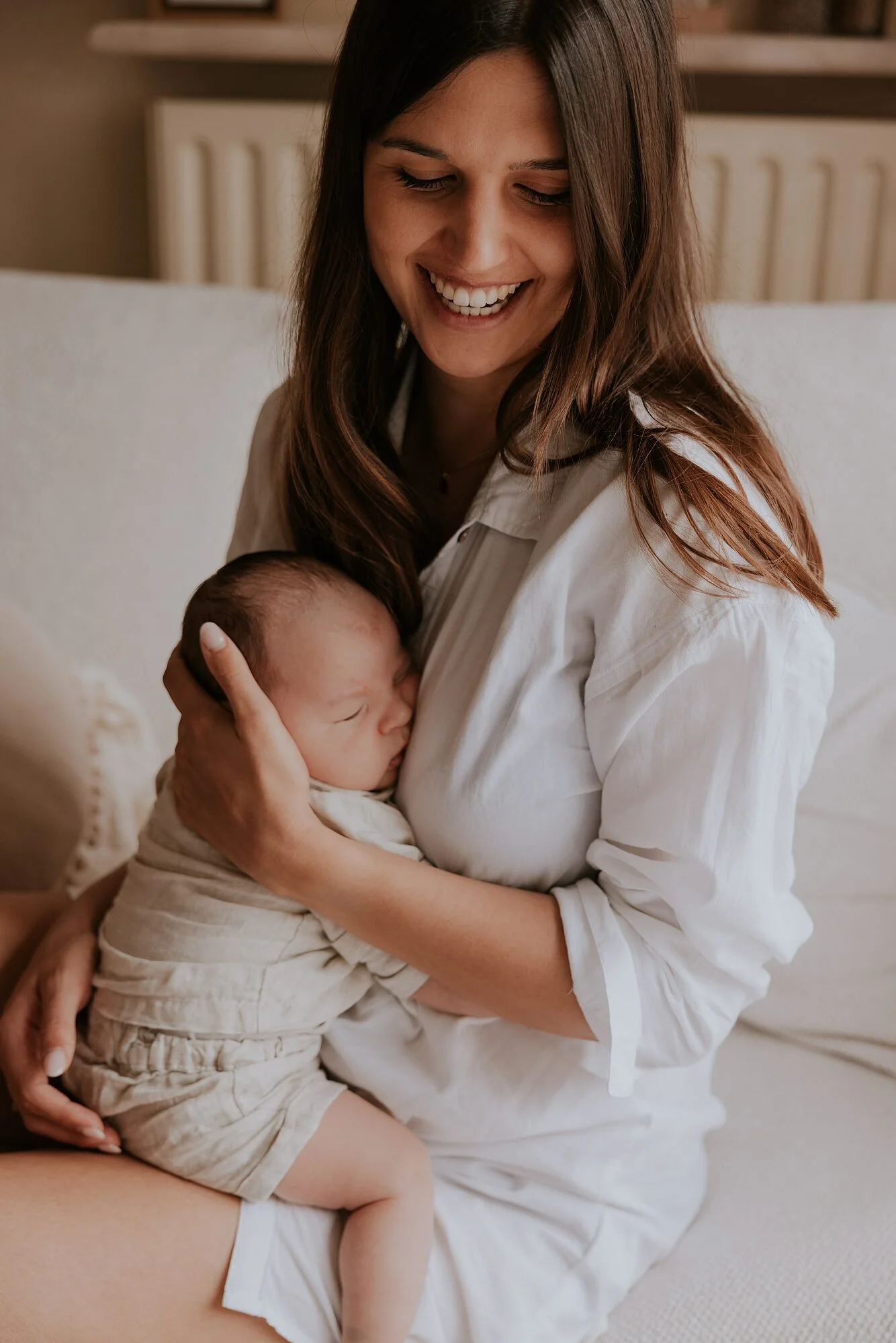 Een vrouw houdt een slapende baby vast terwijl ze op een bed zit, beiden lachen en lijken gelukkig.