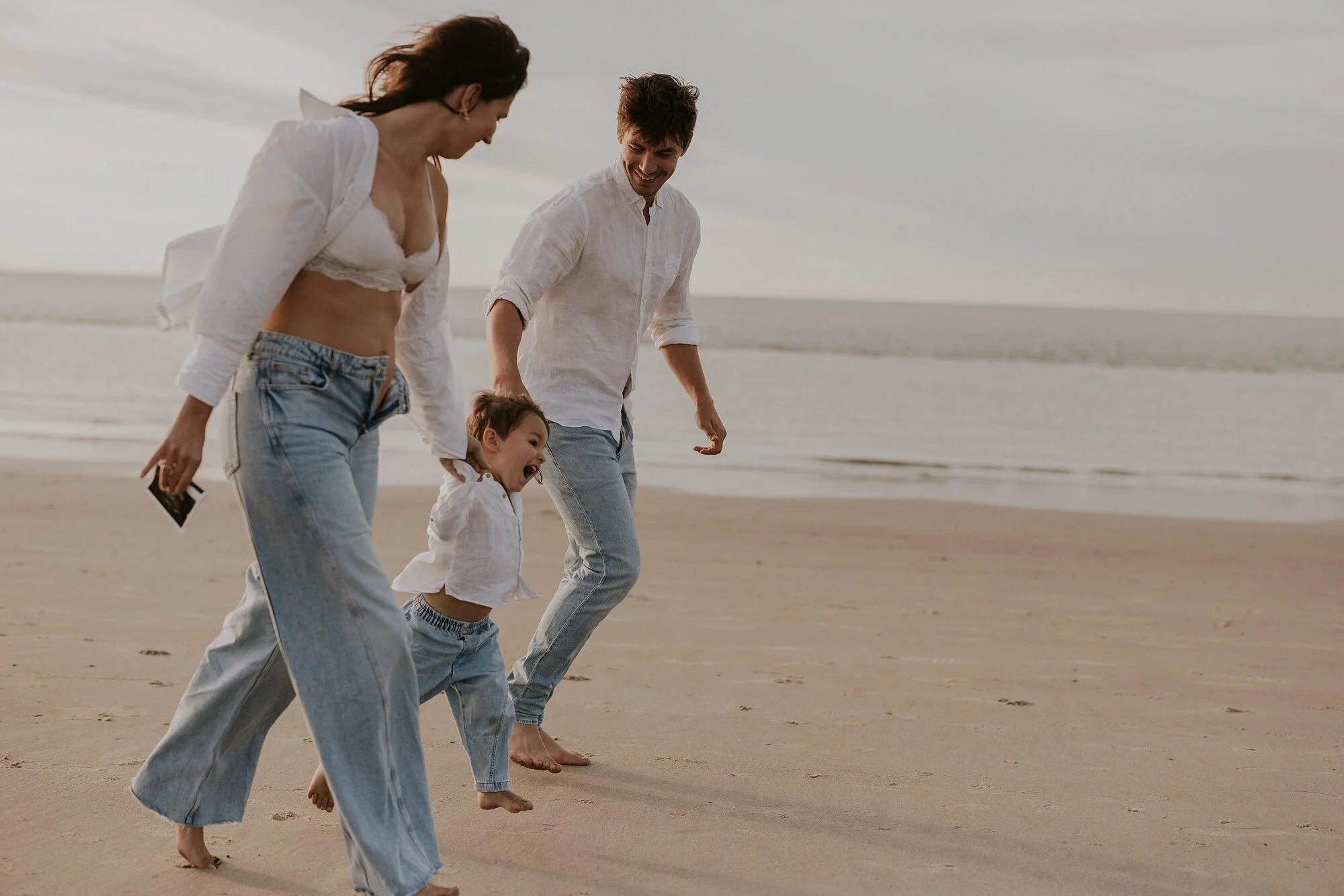 Een gezin met een moeder, vader en zoon lopen blootsvoets langs de oceaan op het strand, lachend en genietend van de tijd samen.