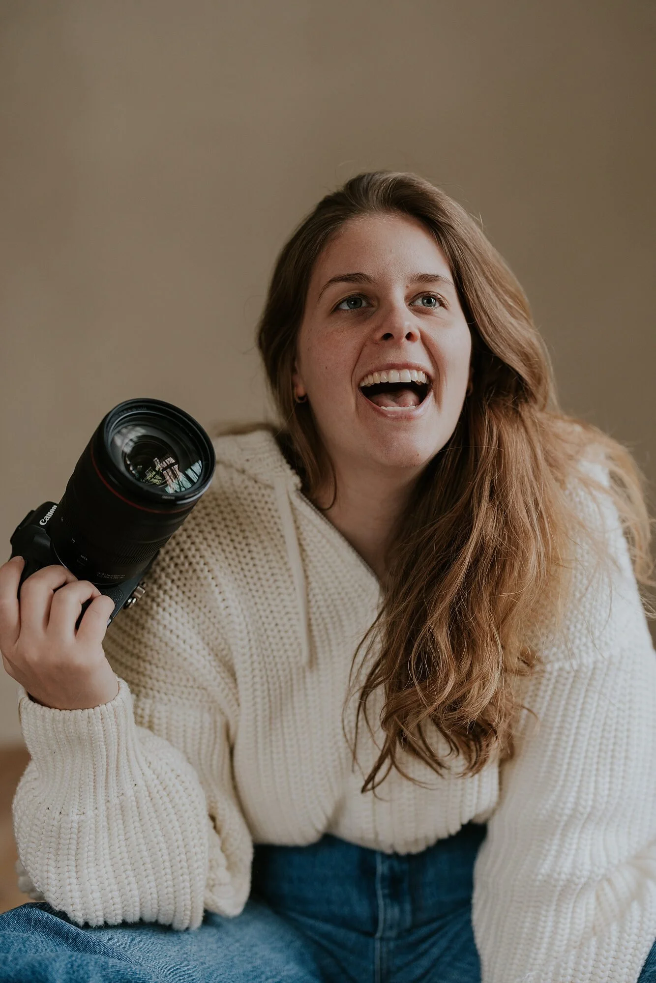 Vrouw met lang, bruin haar, lachend en een camera in haar rechterhand, gekleed in een witte gebreide trui en blauwe spijkerbroek, zittend tegen een beige achtergrond.