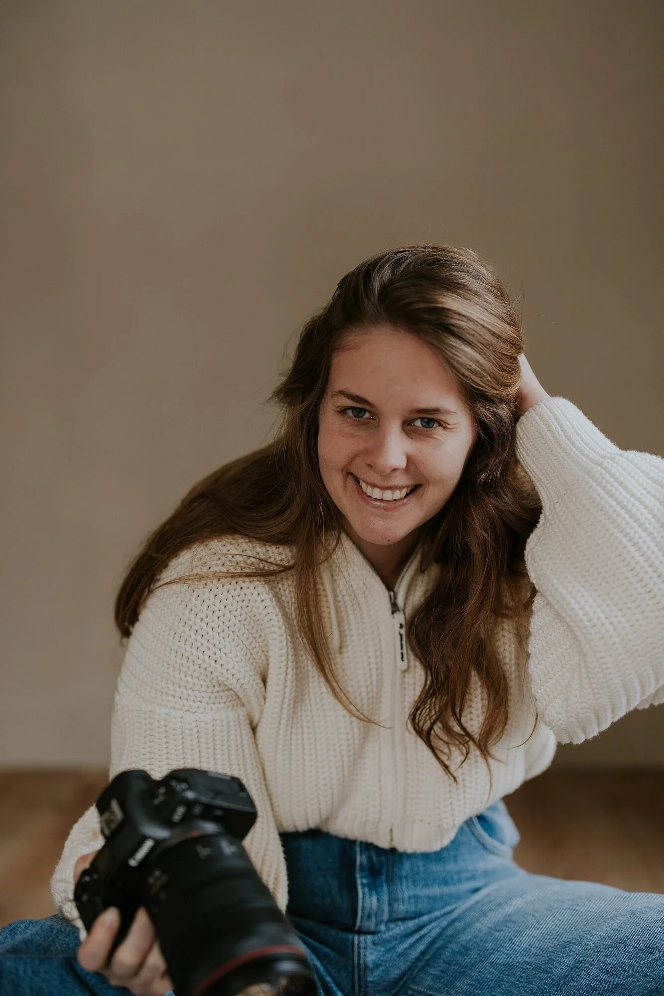 Een lachende jonge vrouw met bruin haar en een witte trui poseert terwijl ze een camera vasthoudt, zittend op de grond, met een effen beige muur op de achtergrond.