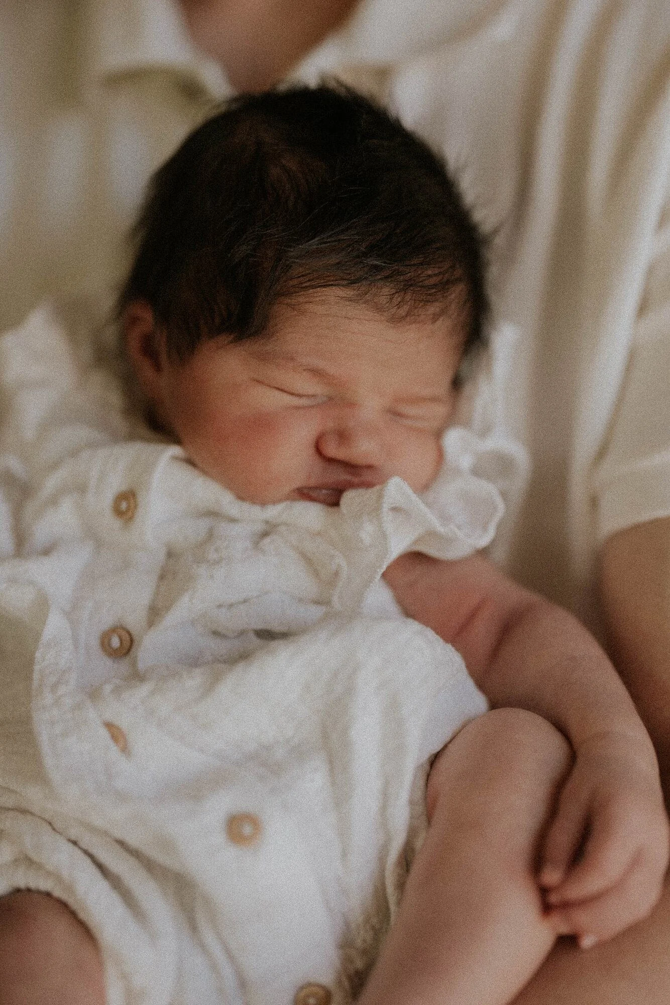 Een slapende baby met donkere, stekelige haar, gekleed in een witte outfit met knoopjes, die wordt vastgehouden door een volwassene.