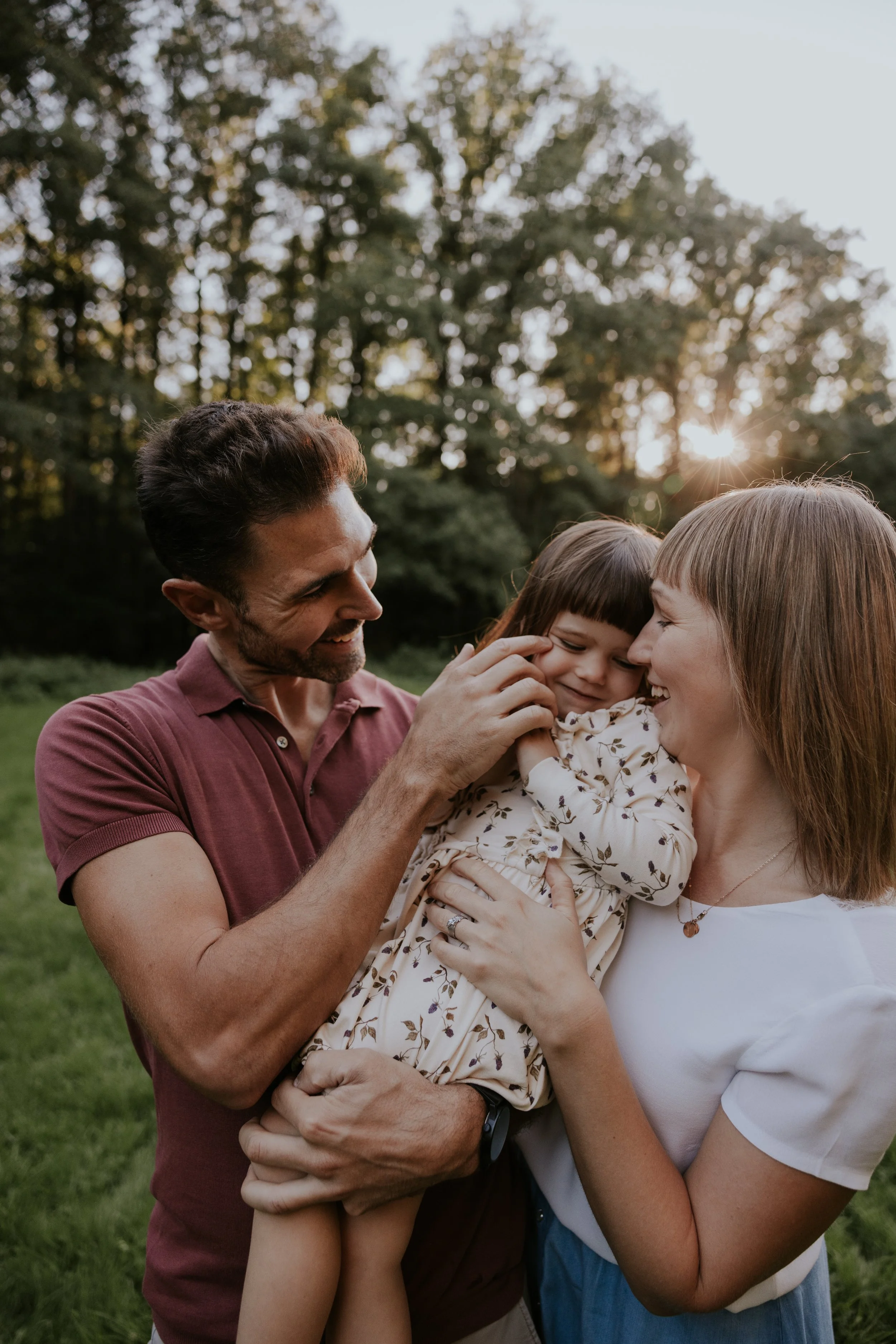 Gezinnetje met twee volwassenen en een kind die lachen in een park tijdens zonsondergang.
