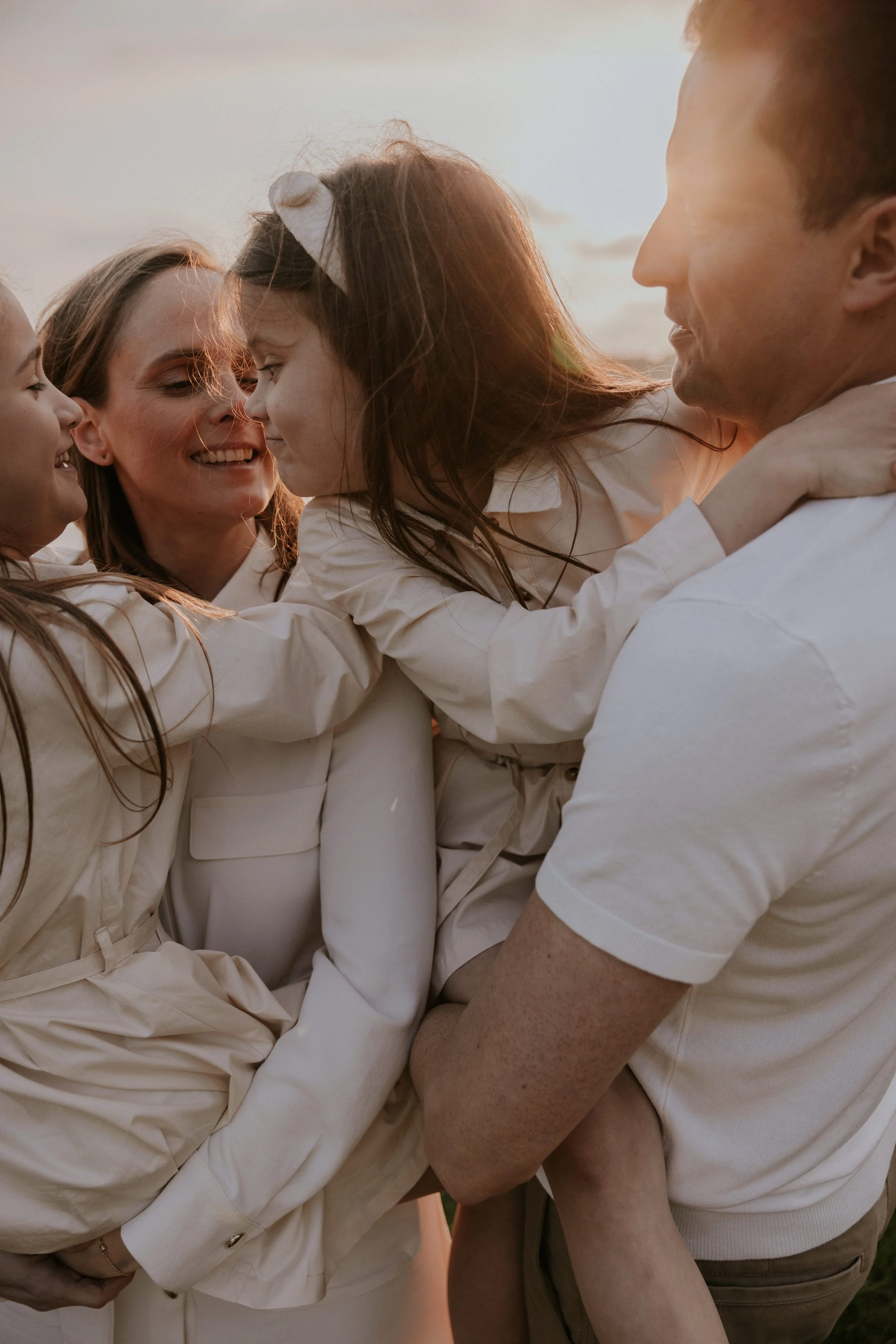 Een gelukkige familie die elkaar omhelst bij zonsondergang, bestaande uit twee volwassenen en twee kinderen, allemaal gekleed in lichte kleding.