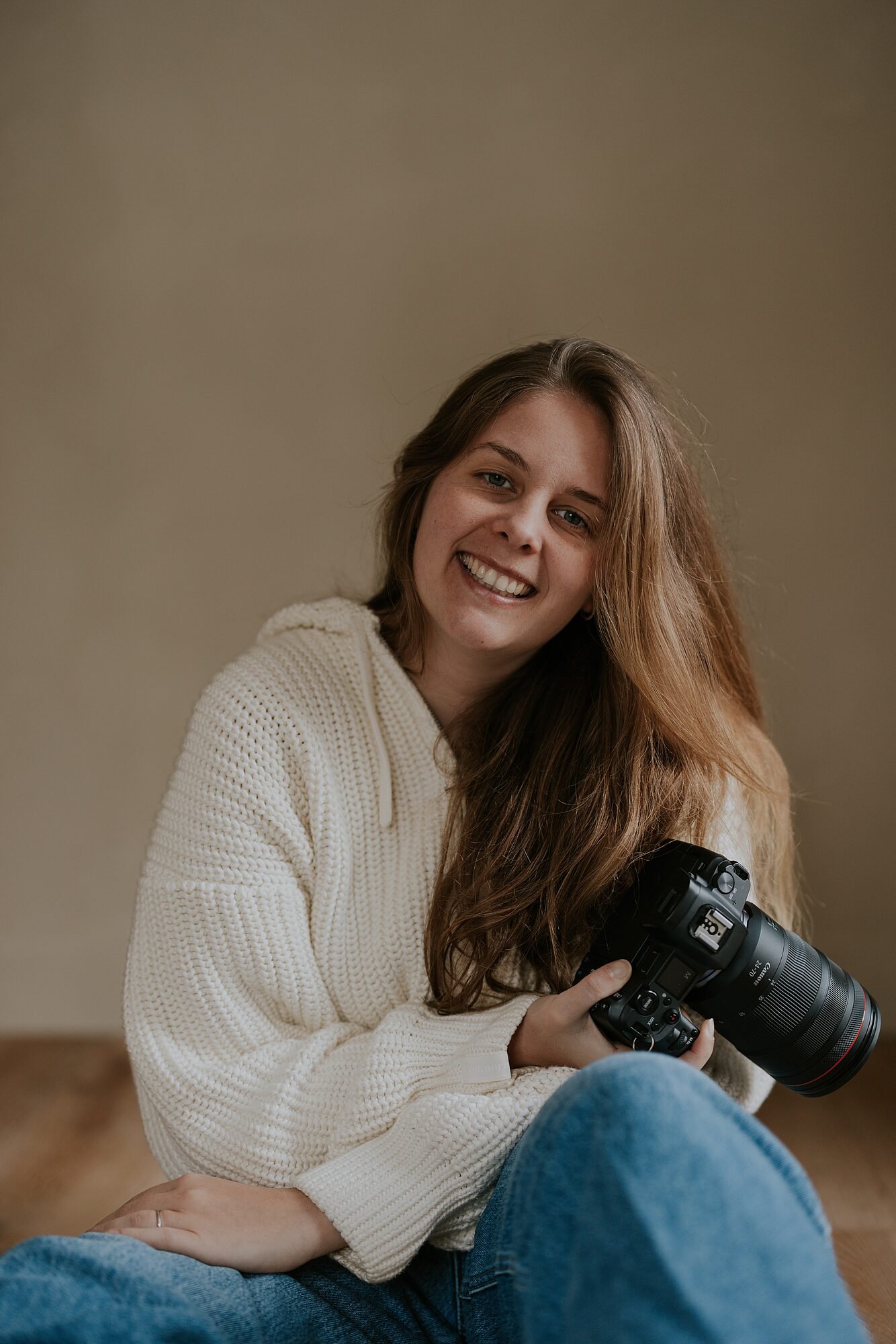 Een glimlachende jonge vrouw met bruin haar die een professionele camera vasthoudt, zittend op de vloer, gekleed in een witte gebreide trui en blauwe jeans.