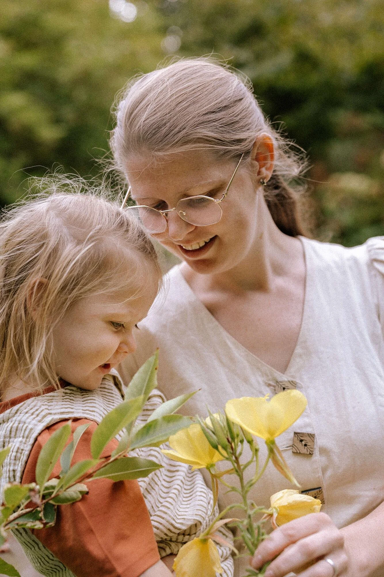 Twee mensen, een volwassene en een kind, naderen een gele bloem, mogelijk in een tuin of park, met een groene achtergrond.