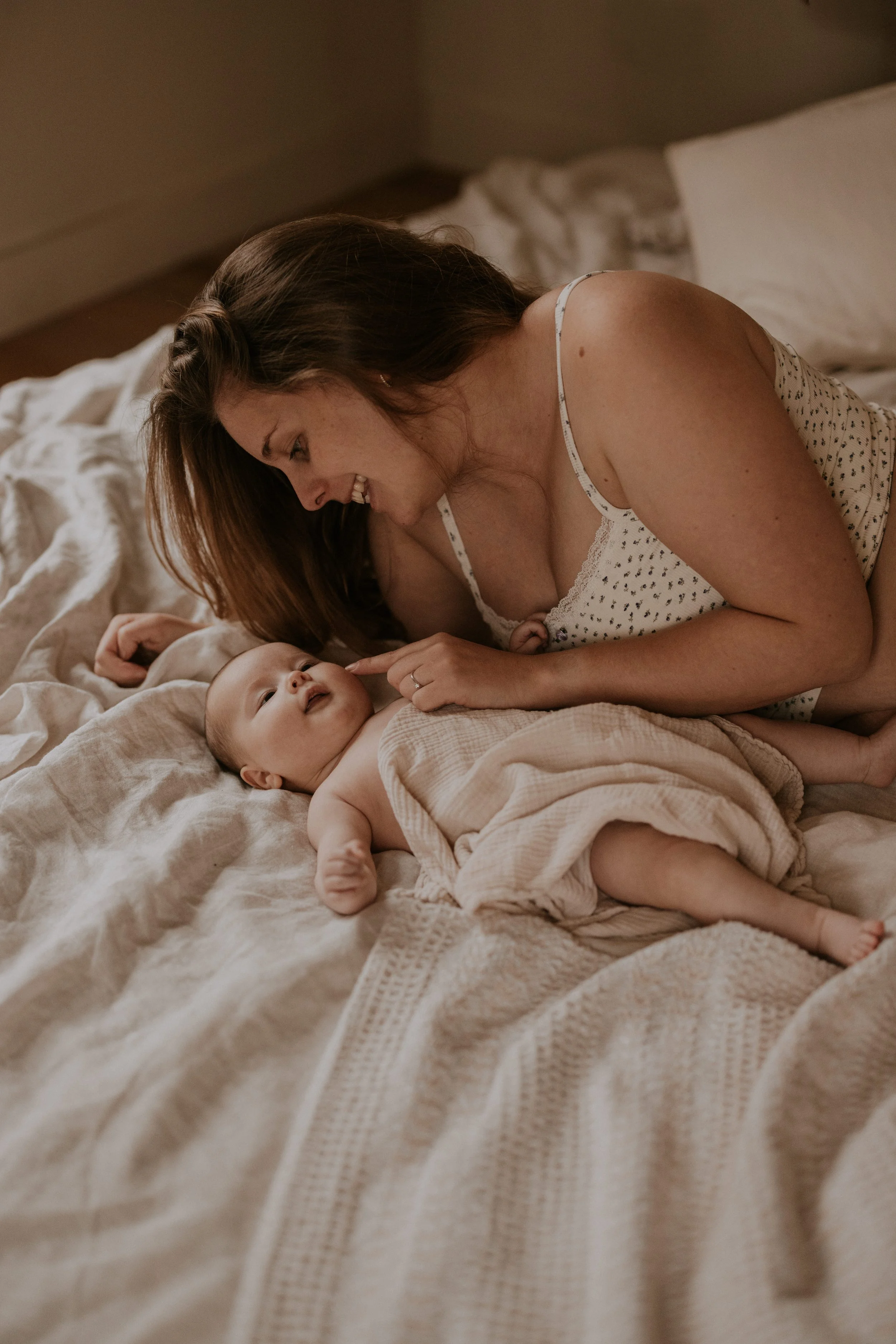 Vrouw met lang bruin haar ligt op een bed, lachend, en speelt met haar baby, die op de rug ligt onder een beige deken.